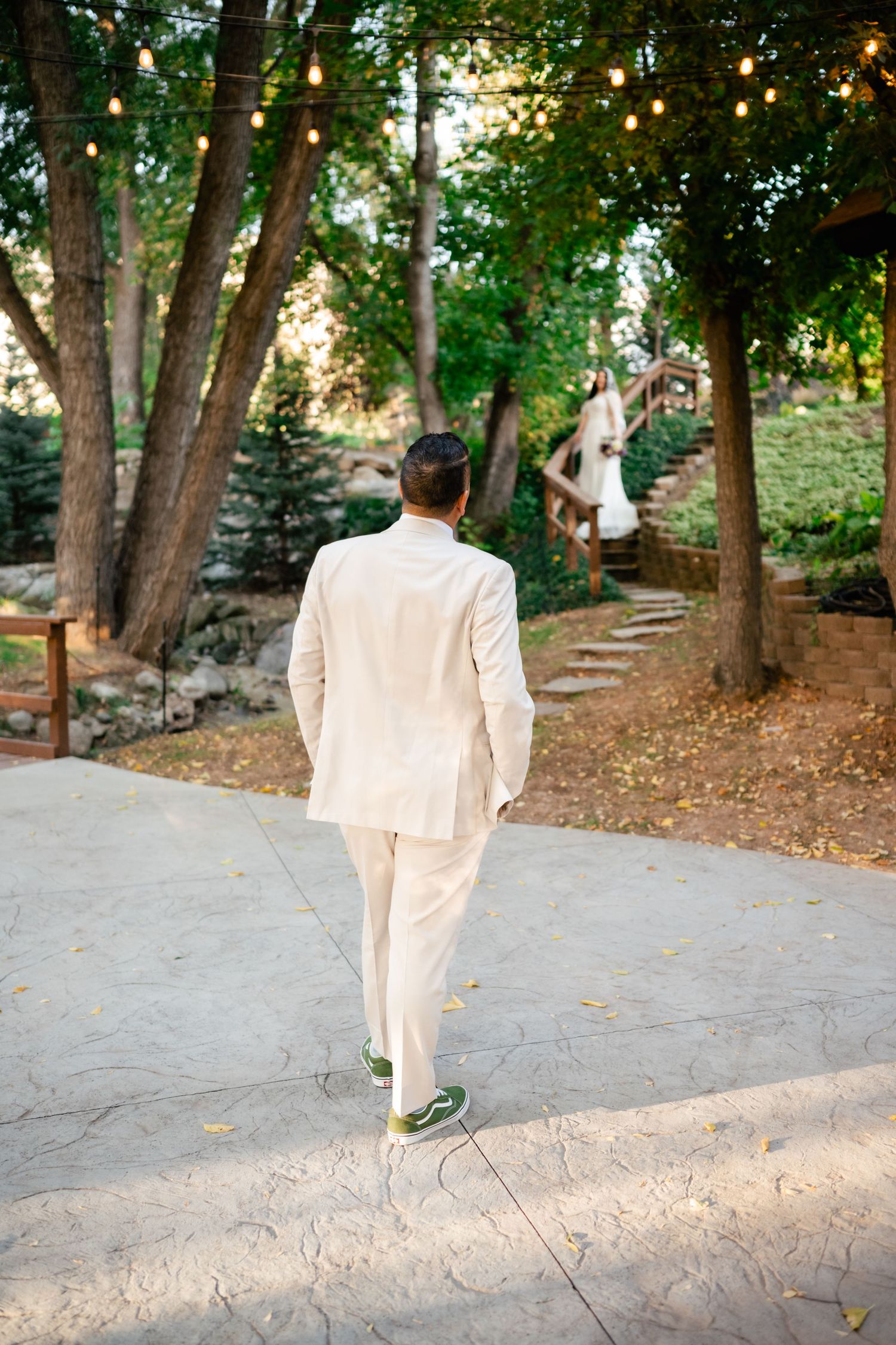 A well-dressed figure in a white suit walks along a path beneath string lights and trees in an outdoor garden setting.