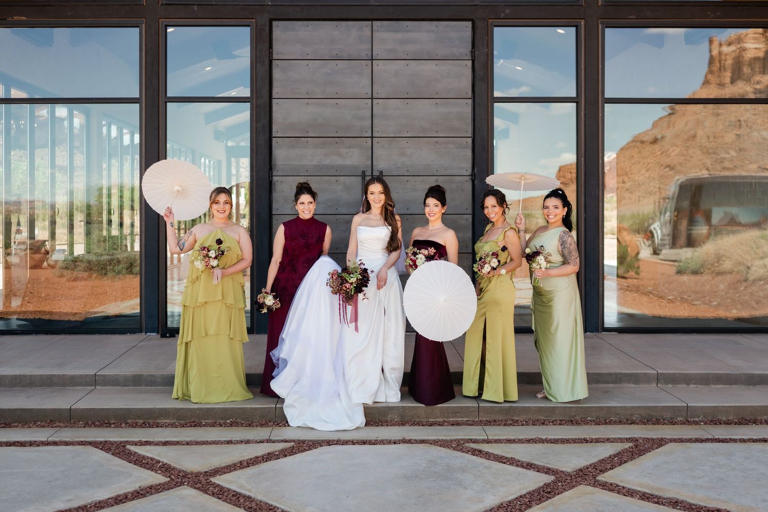 Bride in white gown poses with five bridesmaids in green and burgundy dresses outside a modern venue.