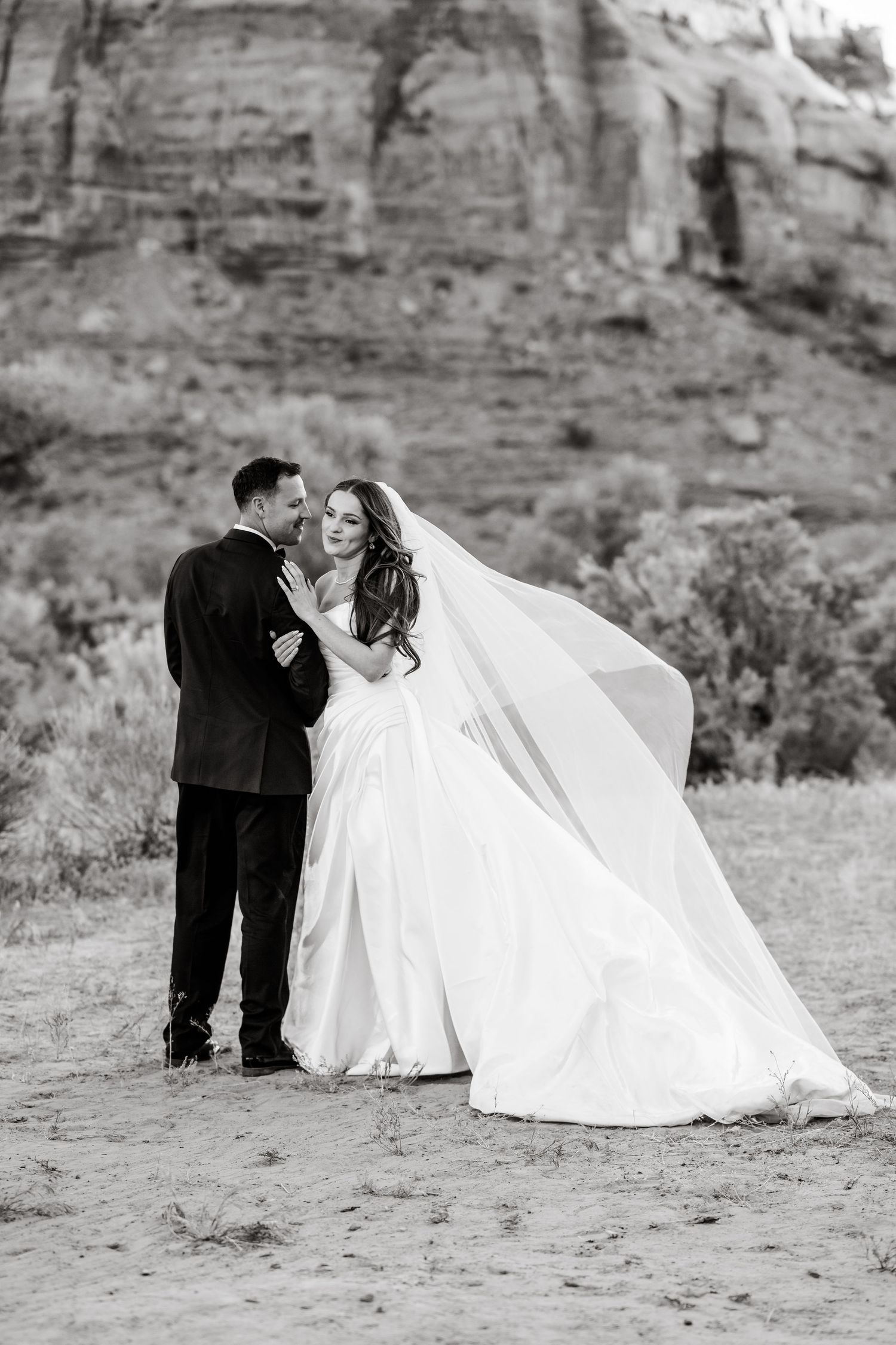 Black and white photo of bride and groom in desert, her veil billowing in the wind with dramatic red rock canyon backdrop.