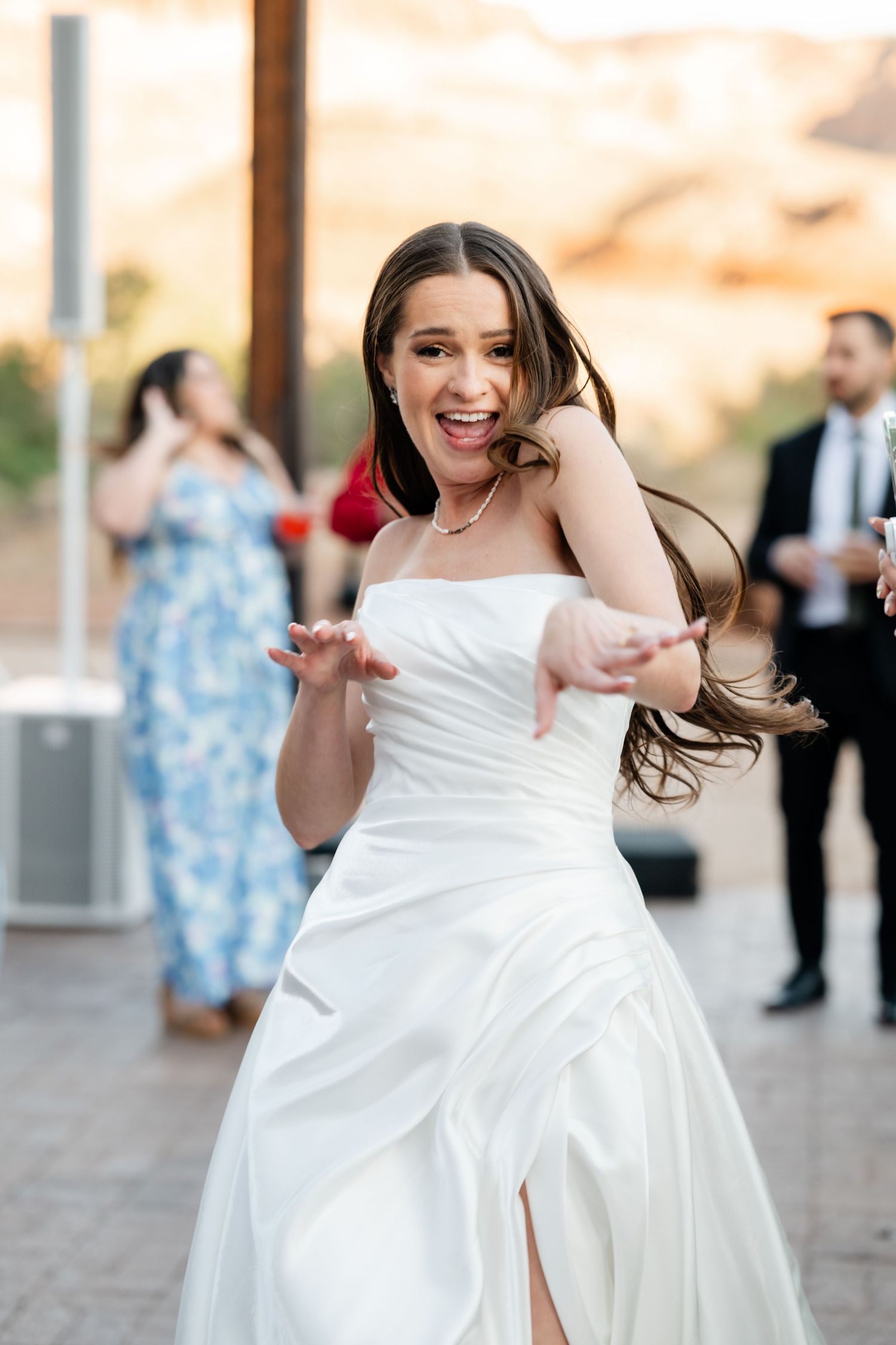 Joyful bride in white strapless gown dancing energetically at outdoor wedding reception with guests in background.