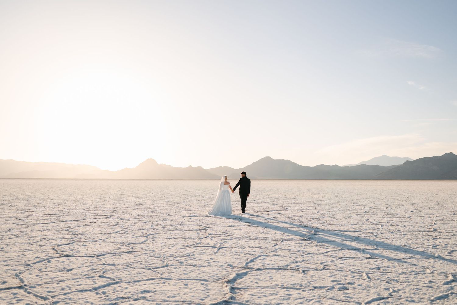 Bride and groom hold hands walking across vast Bonneville Salt Flats at golden sunset with mountains in background.
