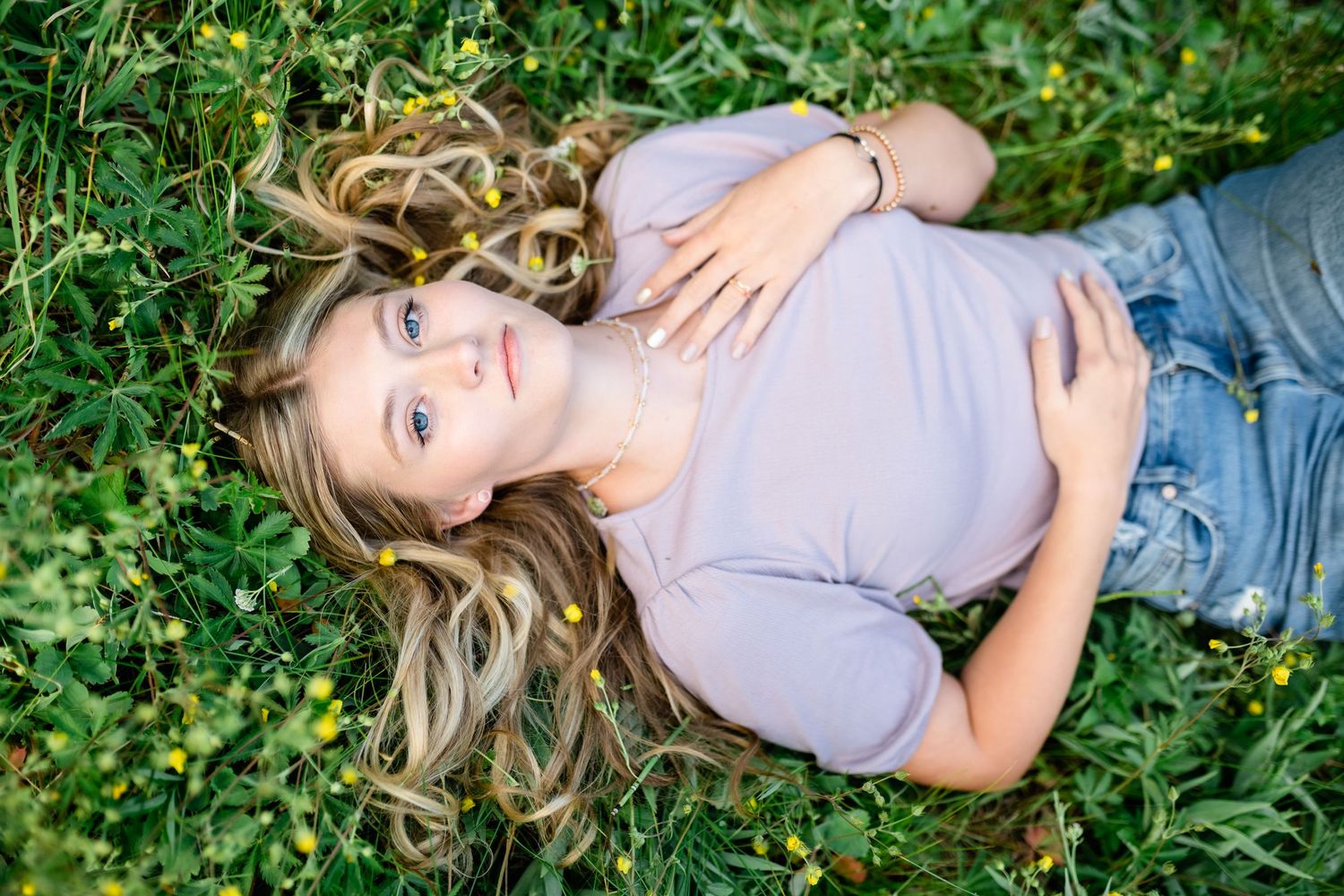 A person lies in a green garden setting wearing a gray top and jeans while surrounded by lush foliage.