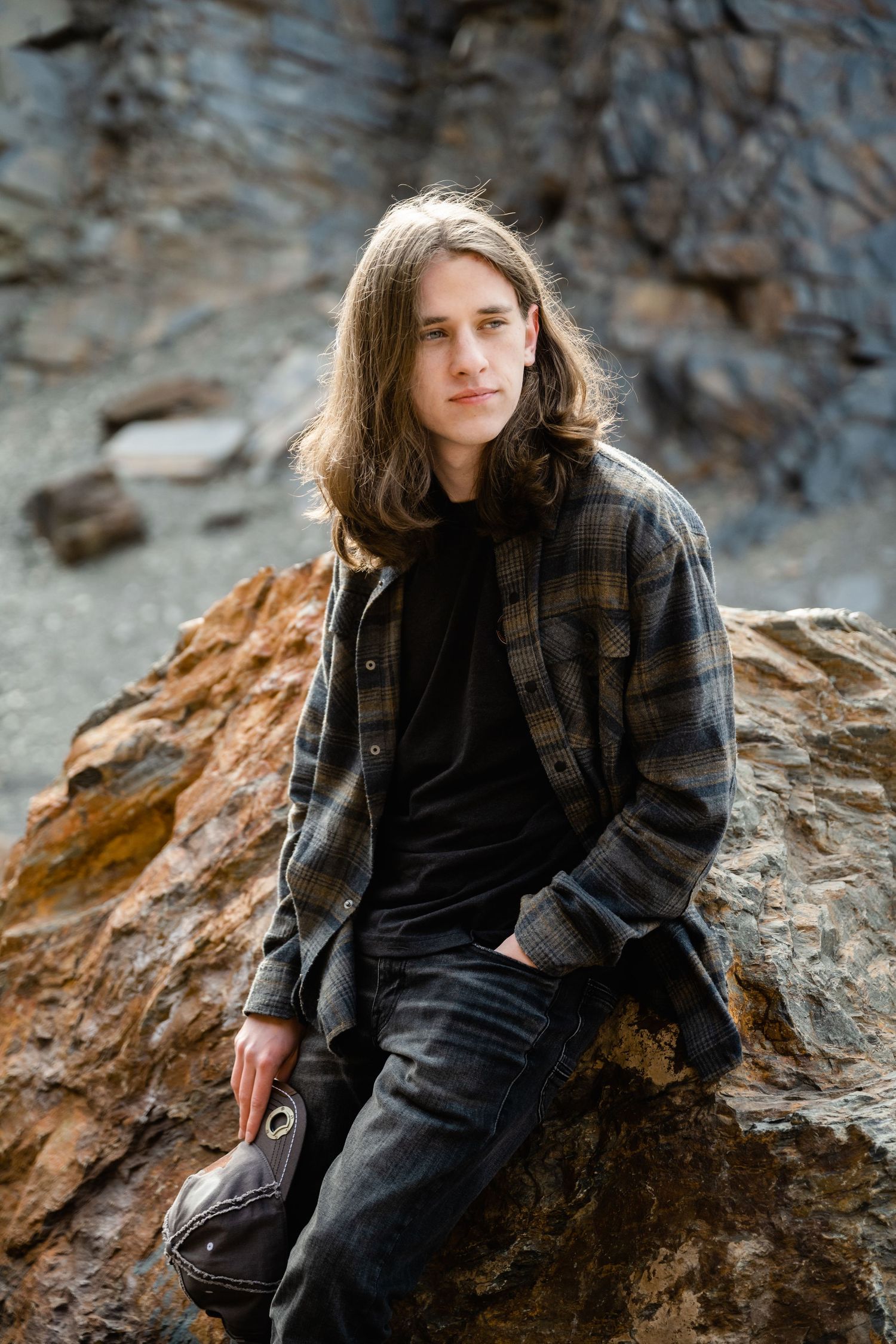 Person in dark winter clothing poses thoughtfully against rocky cliff face in moody outdoor portrait series.