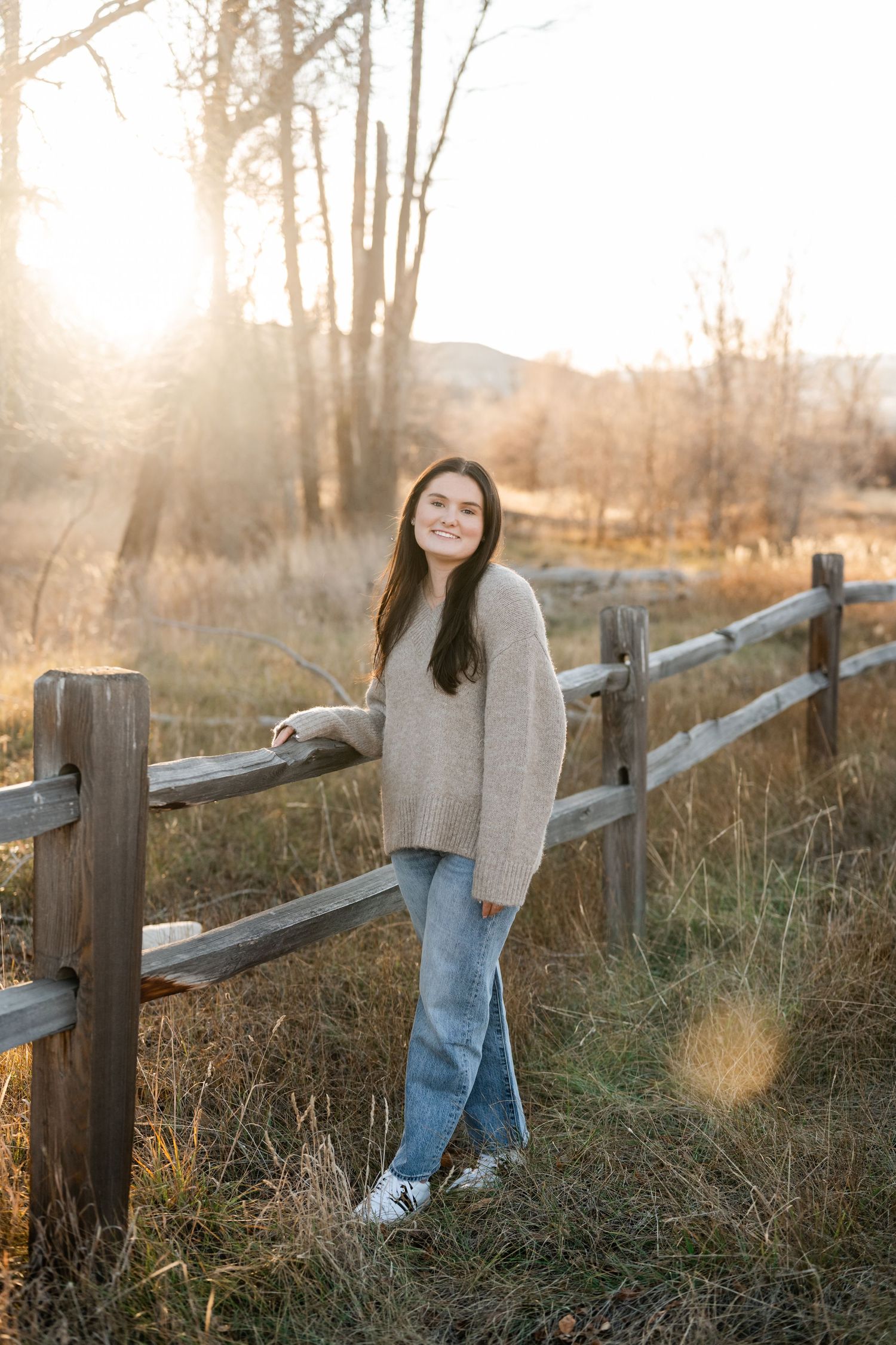 Person in cozy sweater and jeans leans against wooden fence at sunset with golden sunlight filtering through trees.
