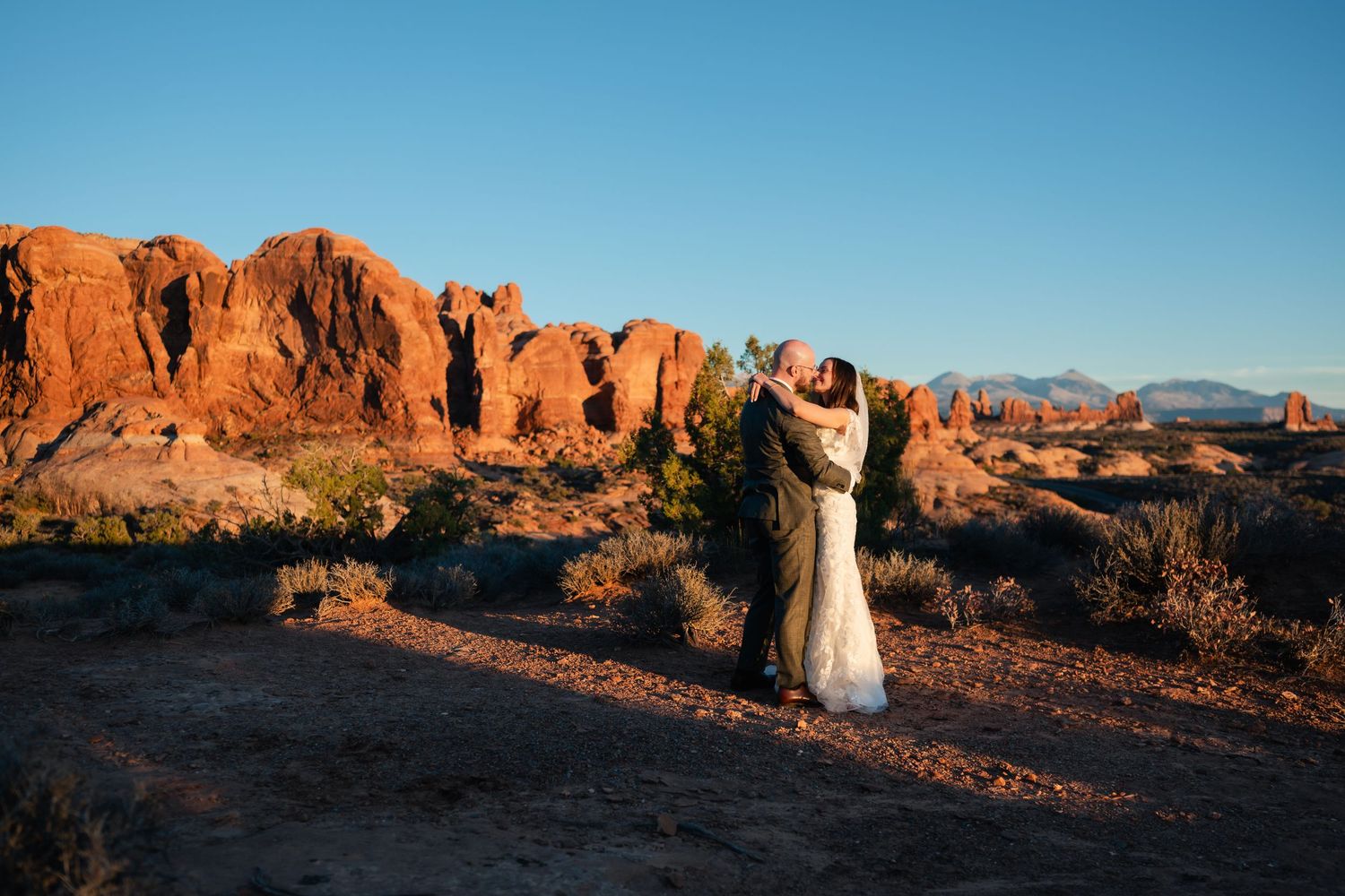A couple embraces during sunset at Arches National Park with red rock formations in the background.