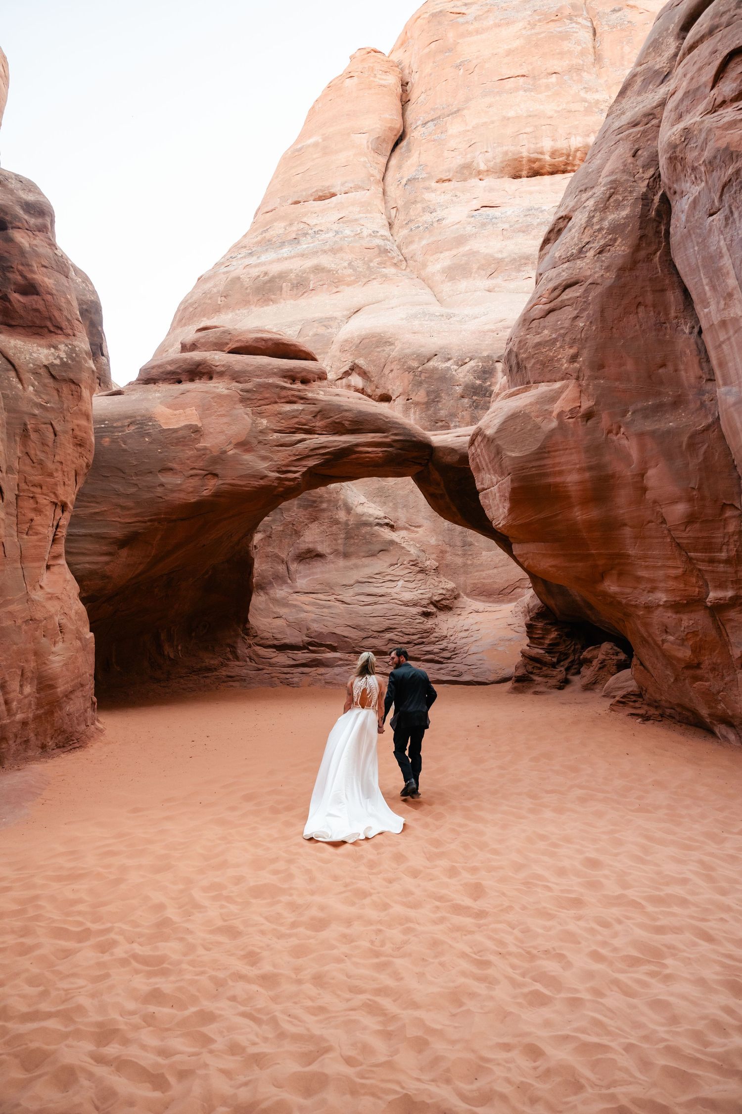A couple in wedding attire embraces under a dramatic red rock arch formation in a desert landscape.