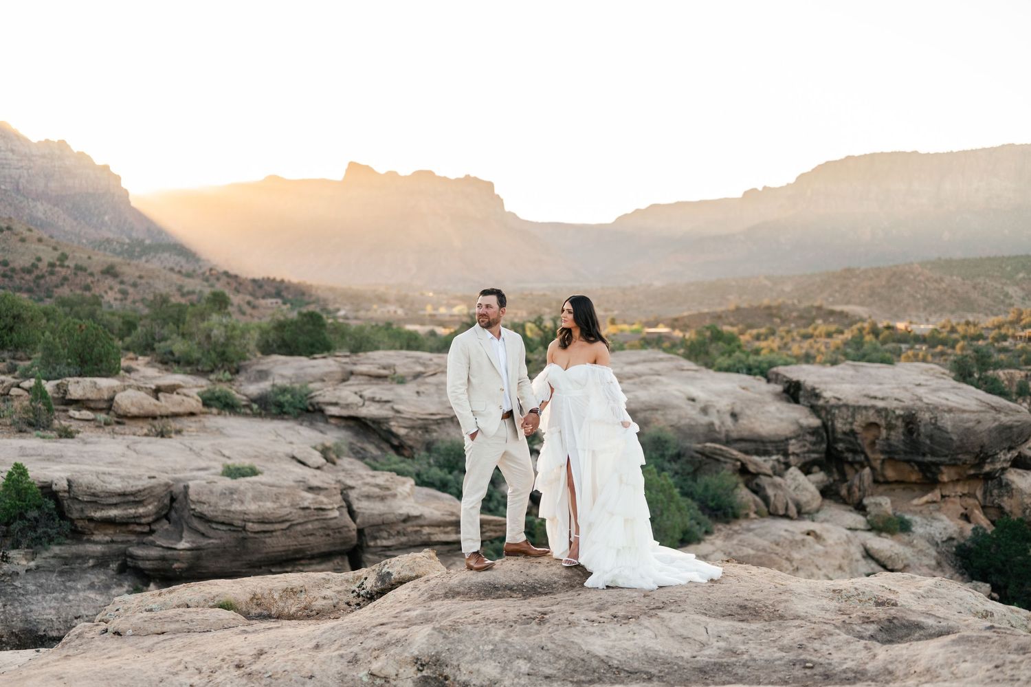 Zion destination photographer. A couple in wedding attire poses on scenic desert rocks during sunset in a mountainous landscape.