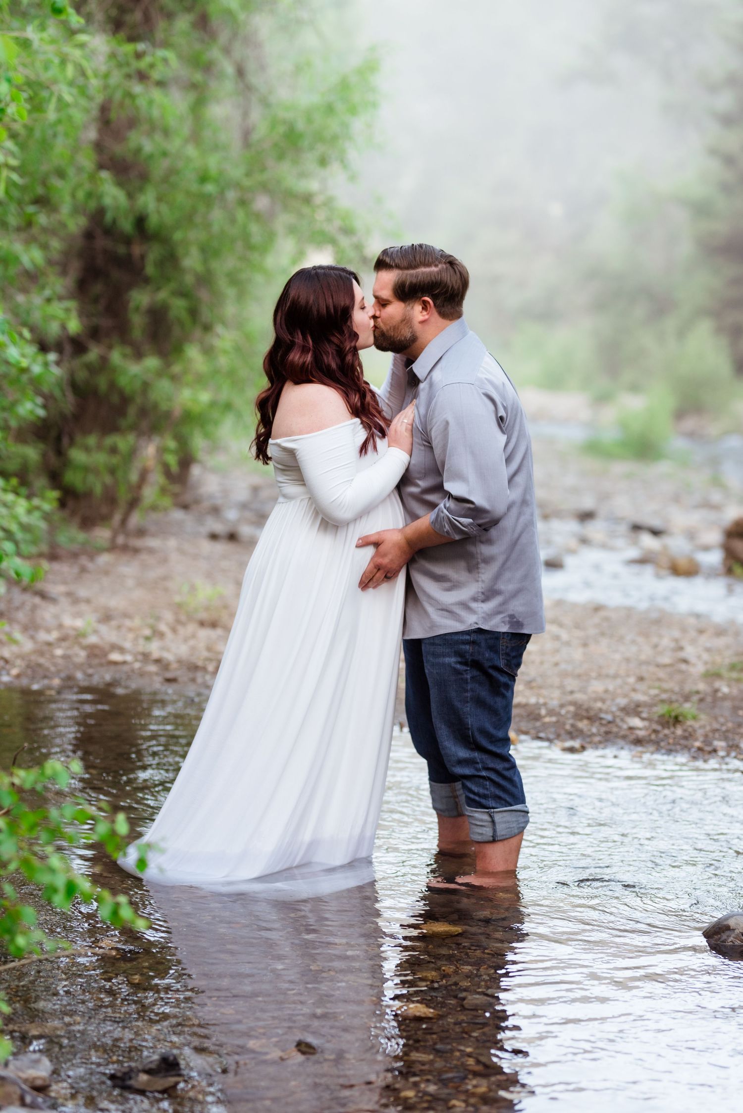 A romantic couple shares intimate moments while standing in a creek surrounded by misty forest scenery.