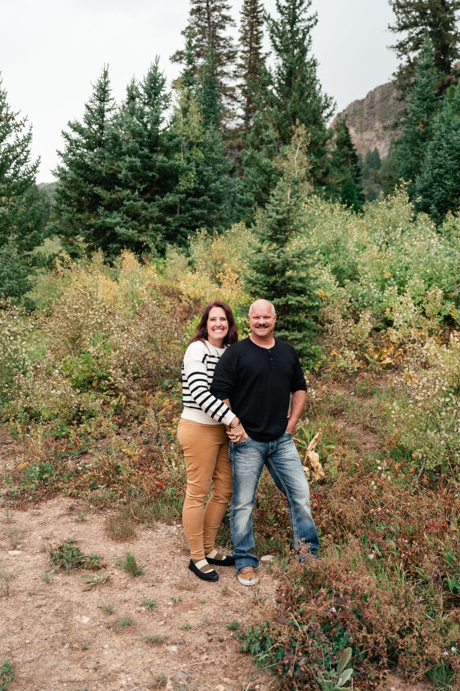 Couple poses together on mountain trail surrounded by evergreen trees and autumn foliage.