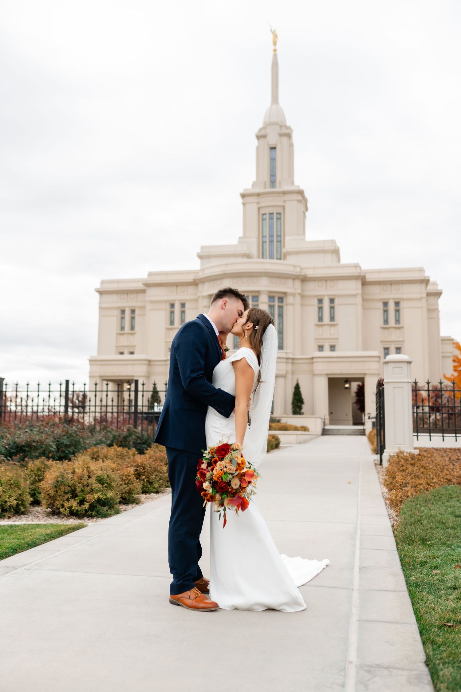Bride and groom kissing in front of an LDS temple on their wedding day, bride holding autumn bouquet.