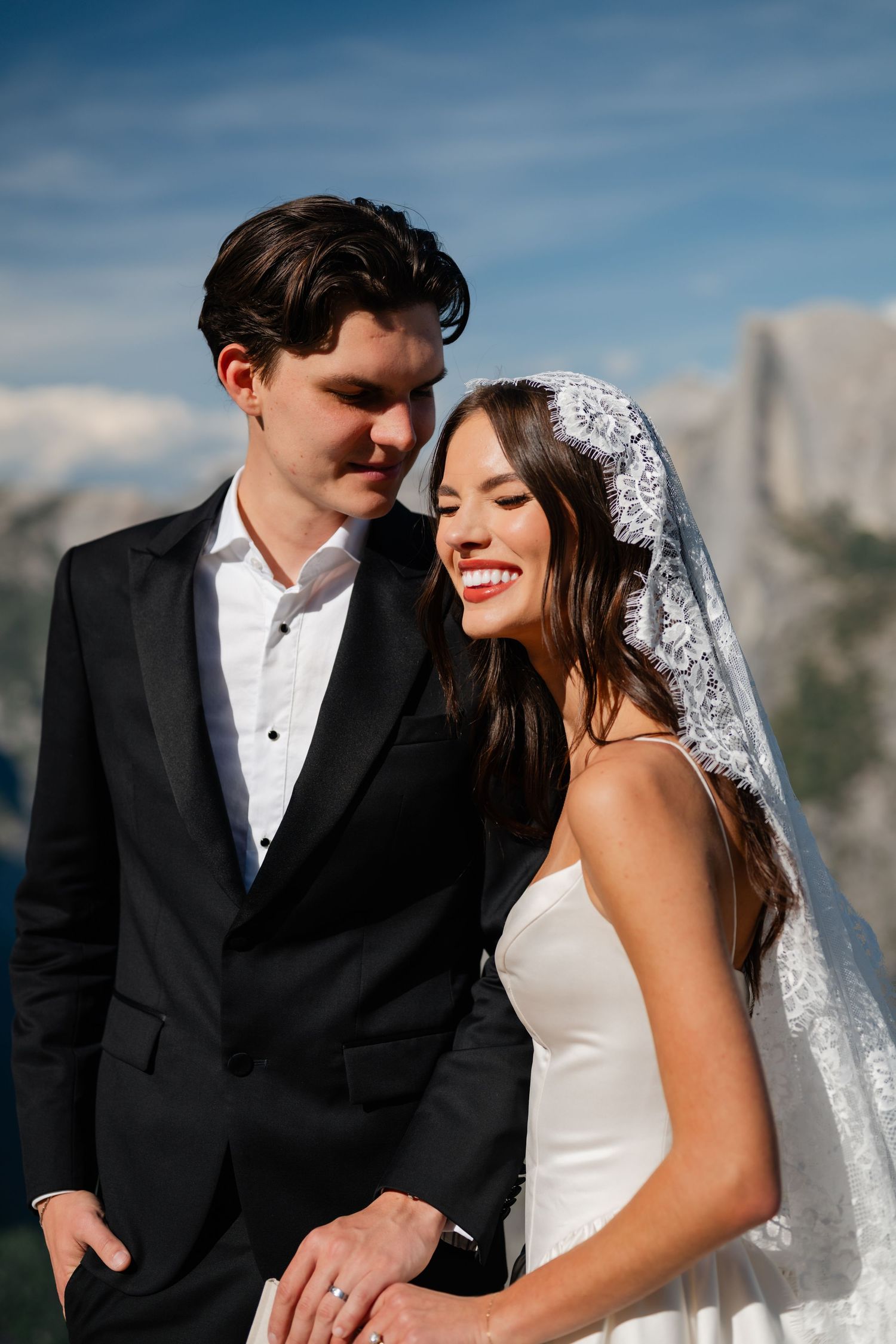Smiling bride in lace veil and groom in black tuxedo pose with Half Dome in Yosemite as backdrop.