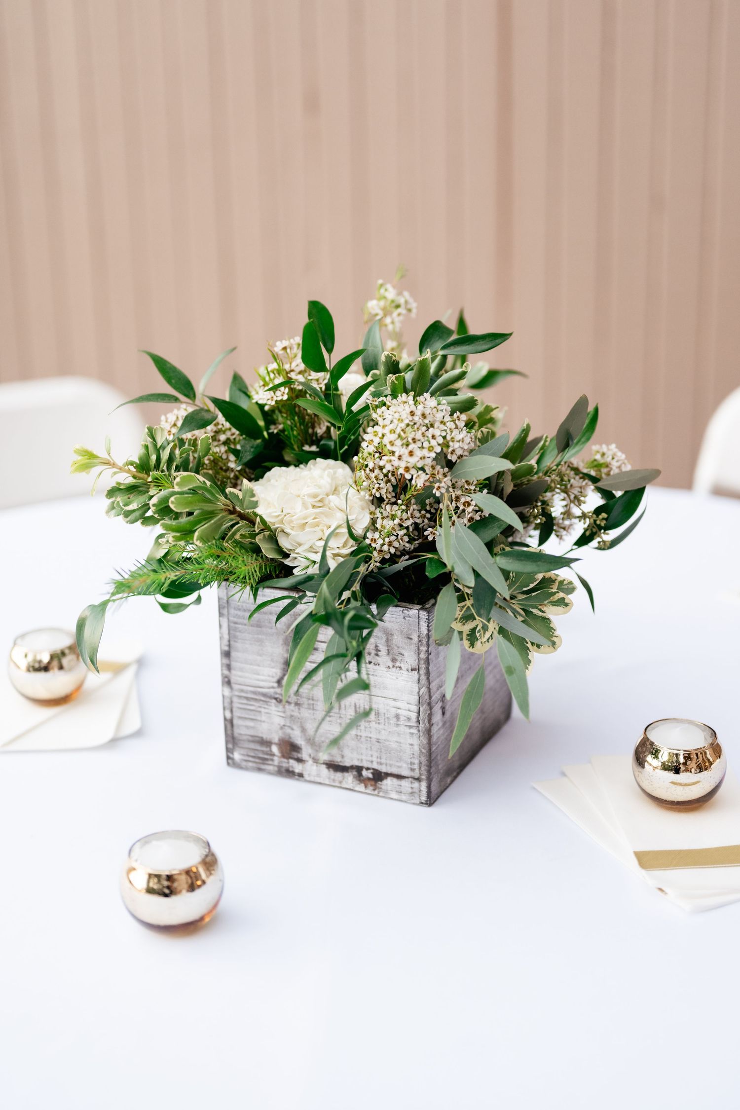 Rustic wooden box centerpiece filled with white flowers and greenery on a clean white surface with metallic accents.