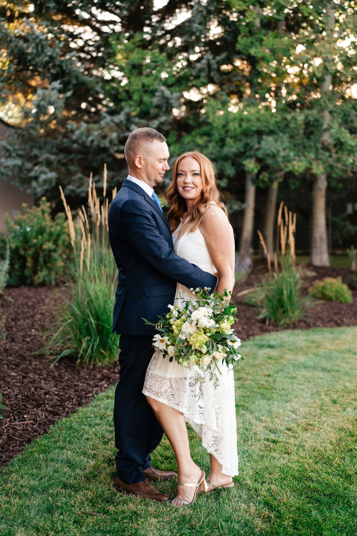 Romantic couple poses together on grassy lawn with white floral bouquet during outdoor photo session.