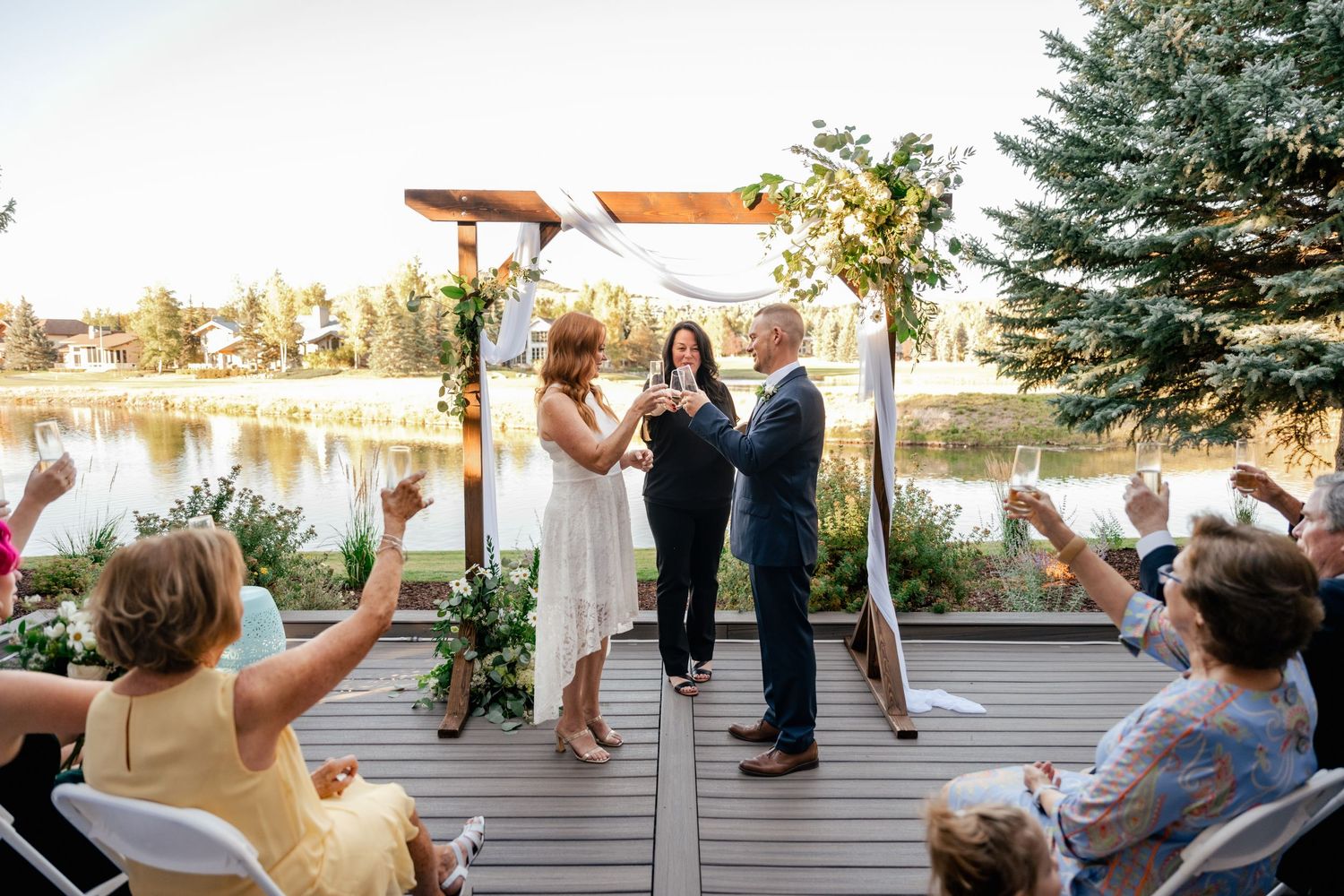Wedding guests seated on white chairs watch ceremony on lakeside deck during golden hour.