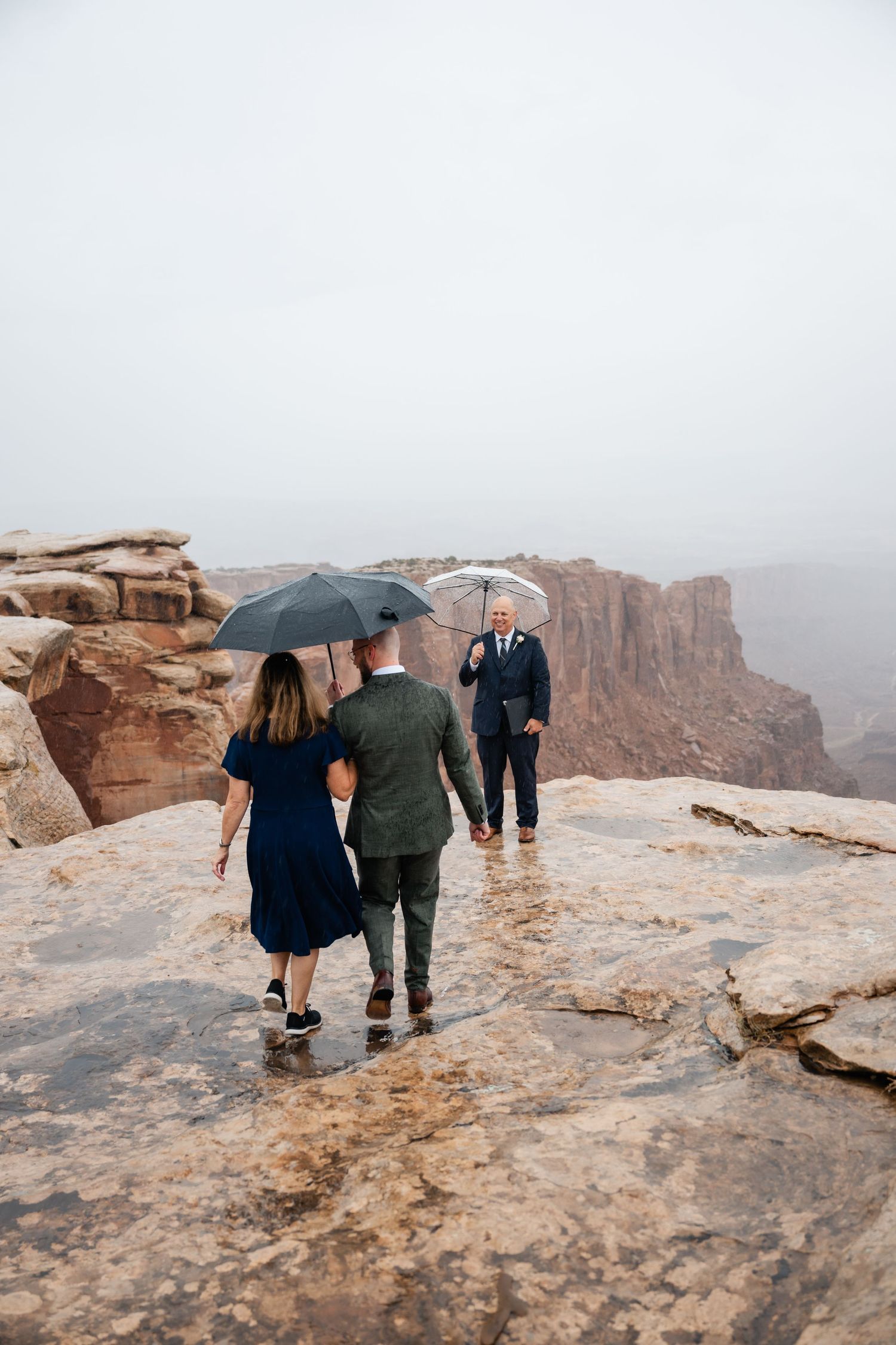 Wedding party walking along cliffside overlook with umbrellas during overcast weather.