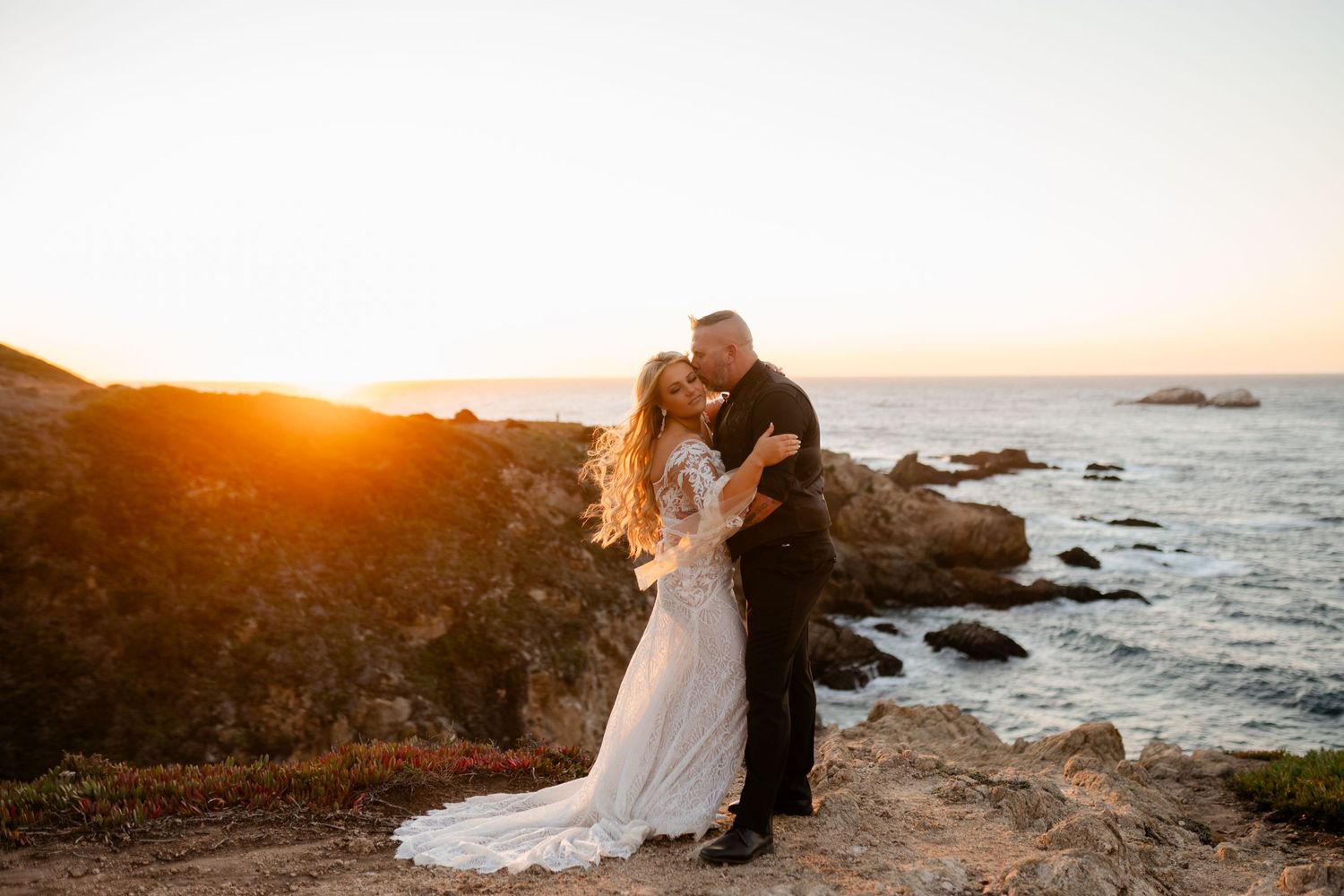 Sunset wedding photo on rocky coastal cliffs with crashing waves below.