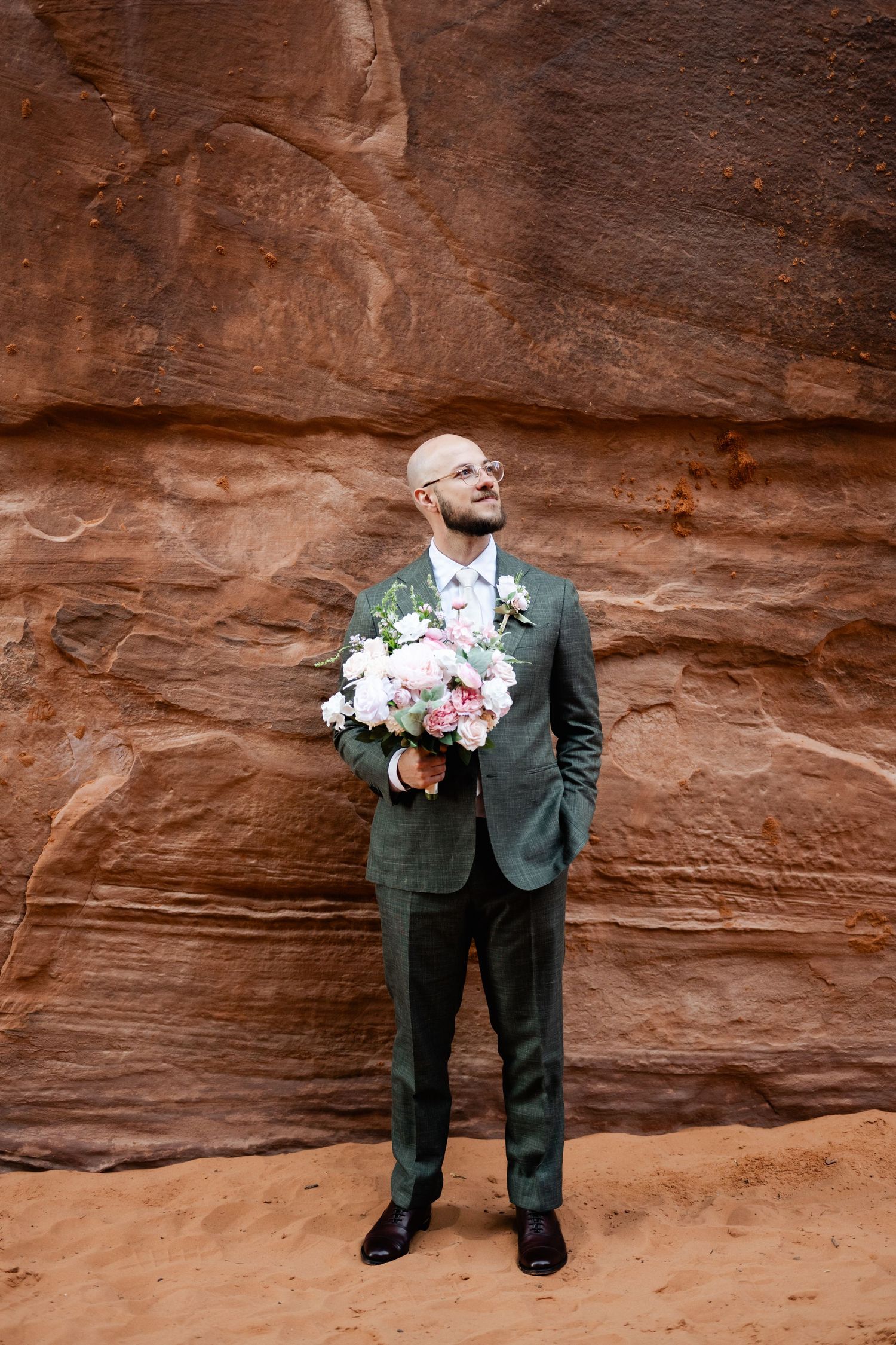 A groom in a grey suit stands against dramatic red rock canyon walls holding a pink and white bouquet.