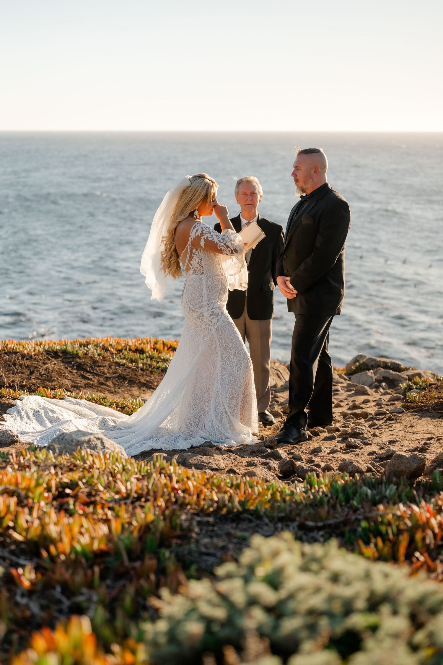 Newlyweds share an intimate moment during their sunset beach wedding ceremony among coastal wildflowers.
