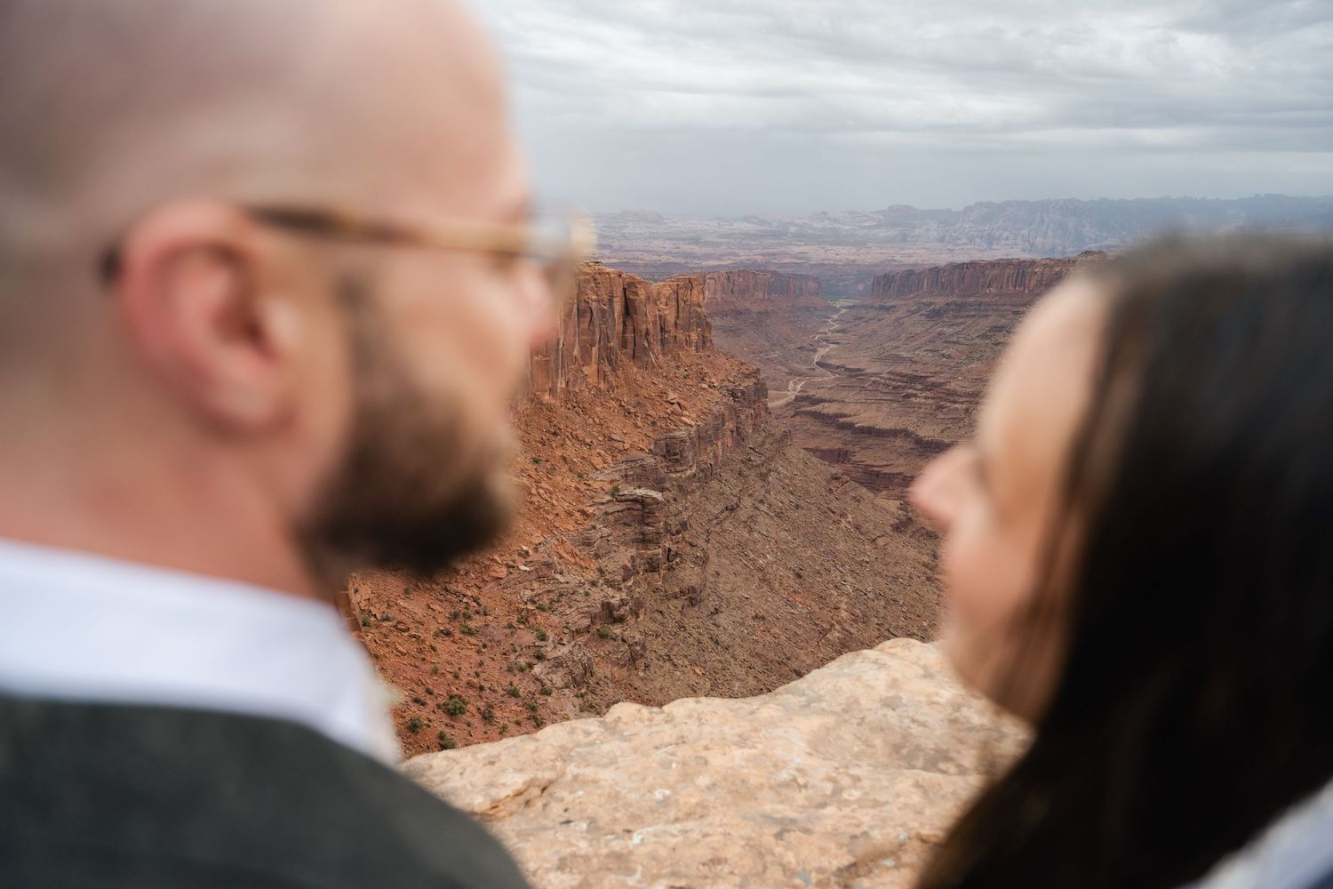 Two people gaze out at a dramatic red rock canyon landscape under cloudy skies in the American Southwest.