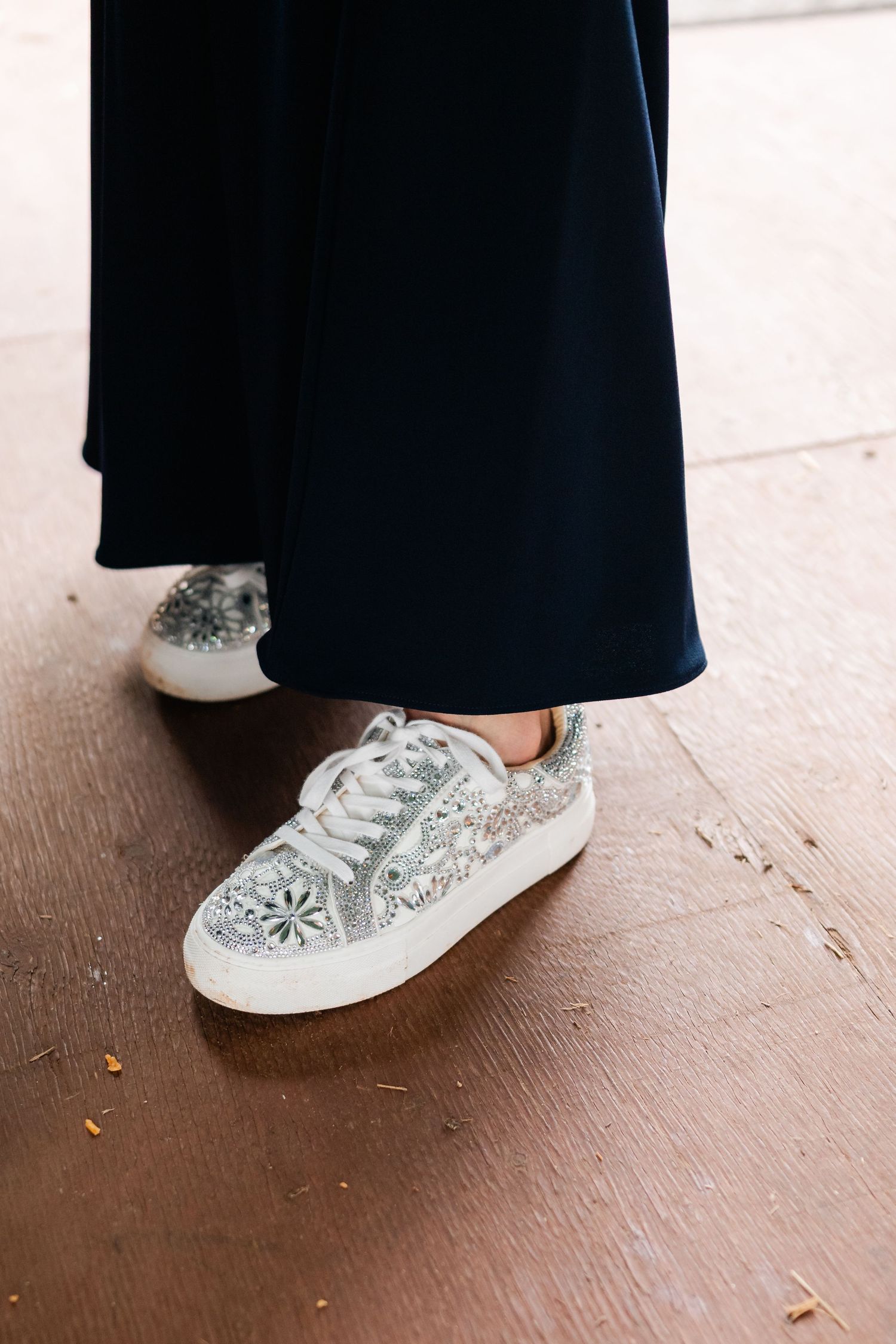 Sparkly silver floral sneakers peek out beneath a long navy dress on a wooden floor.