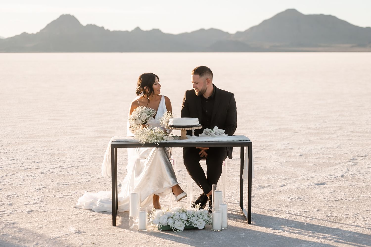 Wedding couple sitting at table on salt flats with mountain landscape in background.