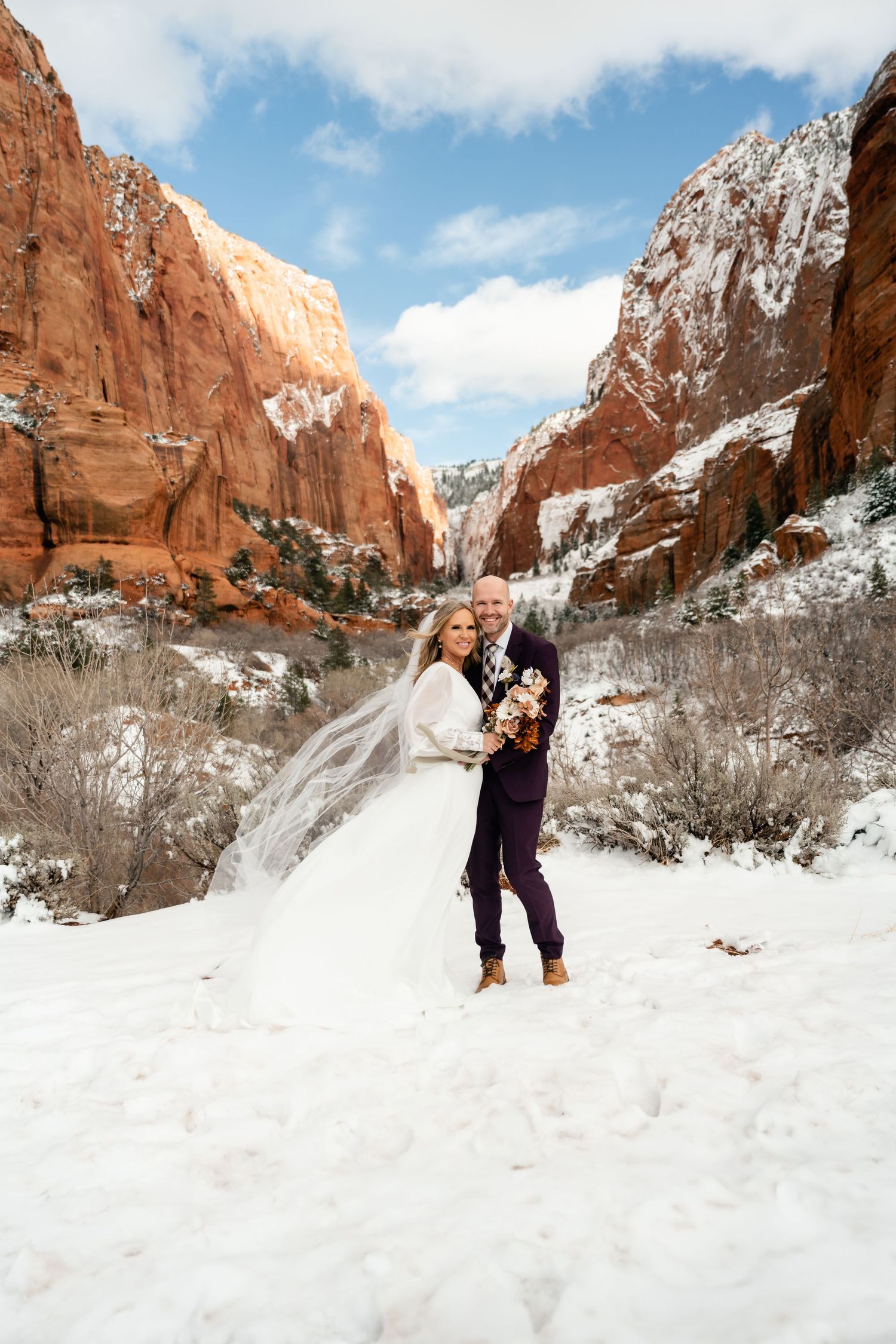 A wedding couple poses in the snow against towering red cliffs in a desert canyon.