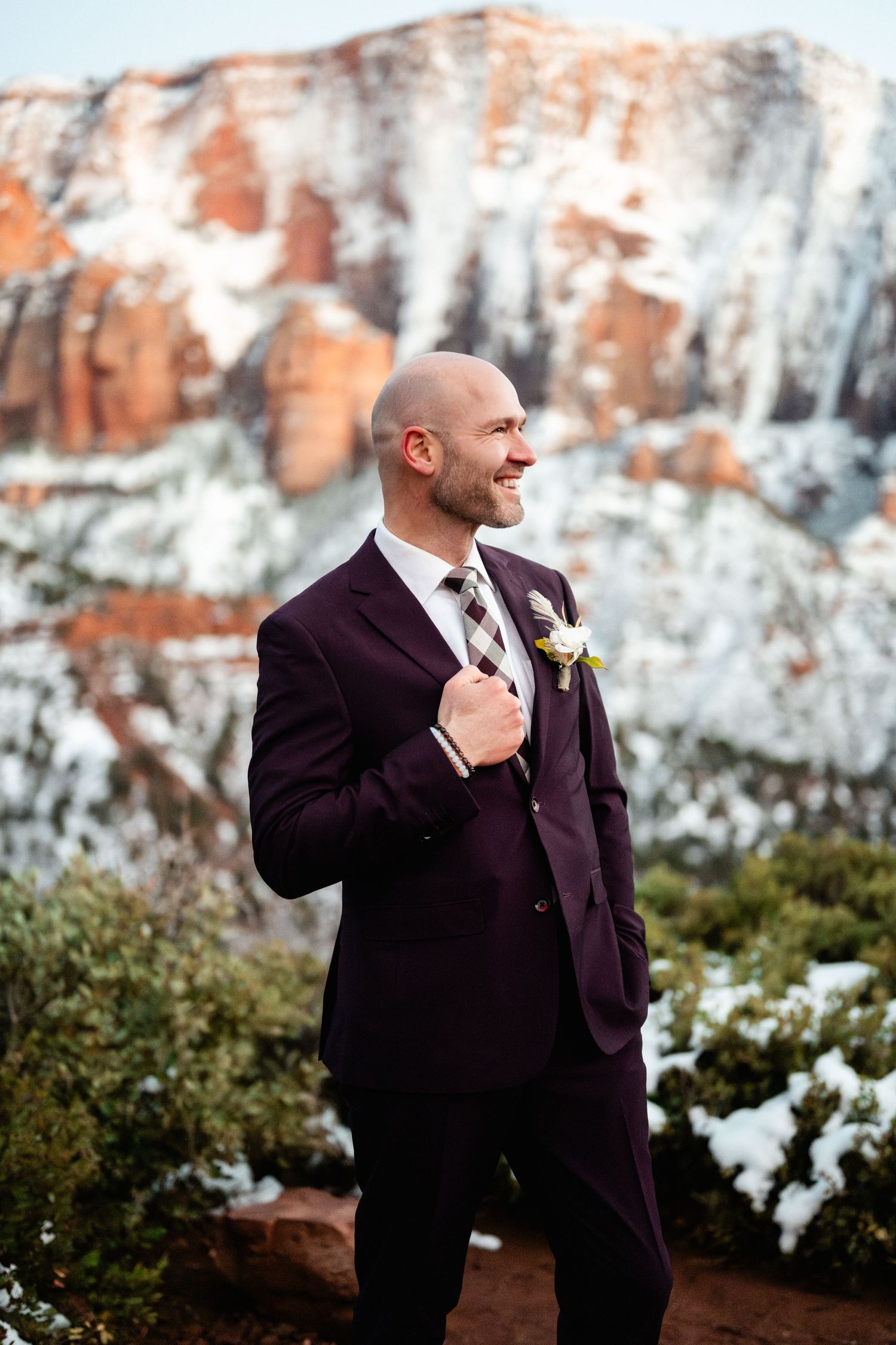 Groom in burgundy suit adjusts tie against snowy red rock cliffs.