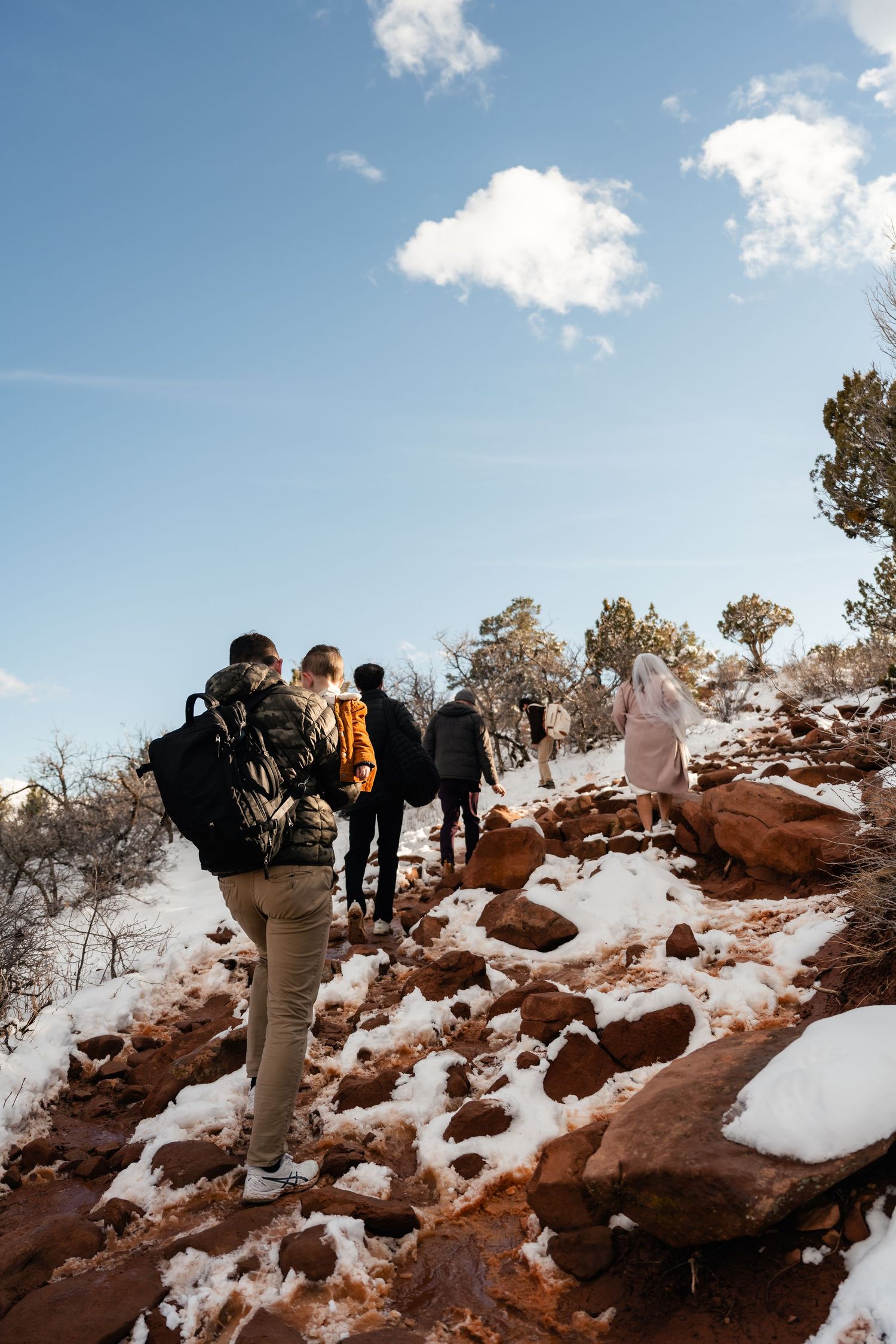 Hikers ascending snowy trail through red rocks under blue winter sky.