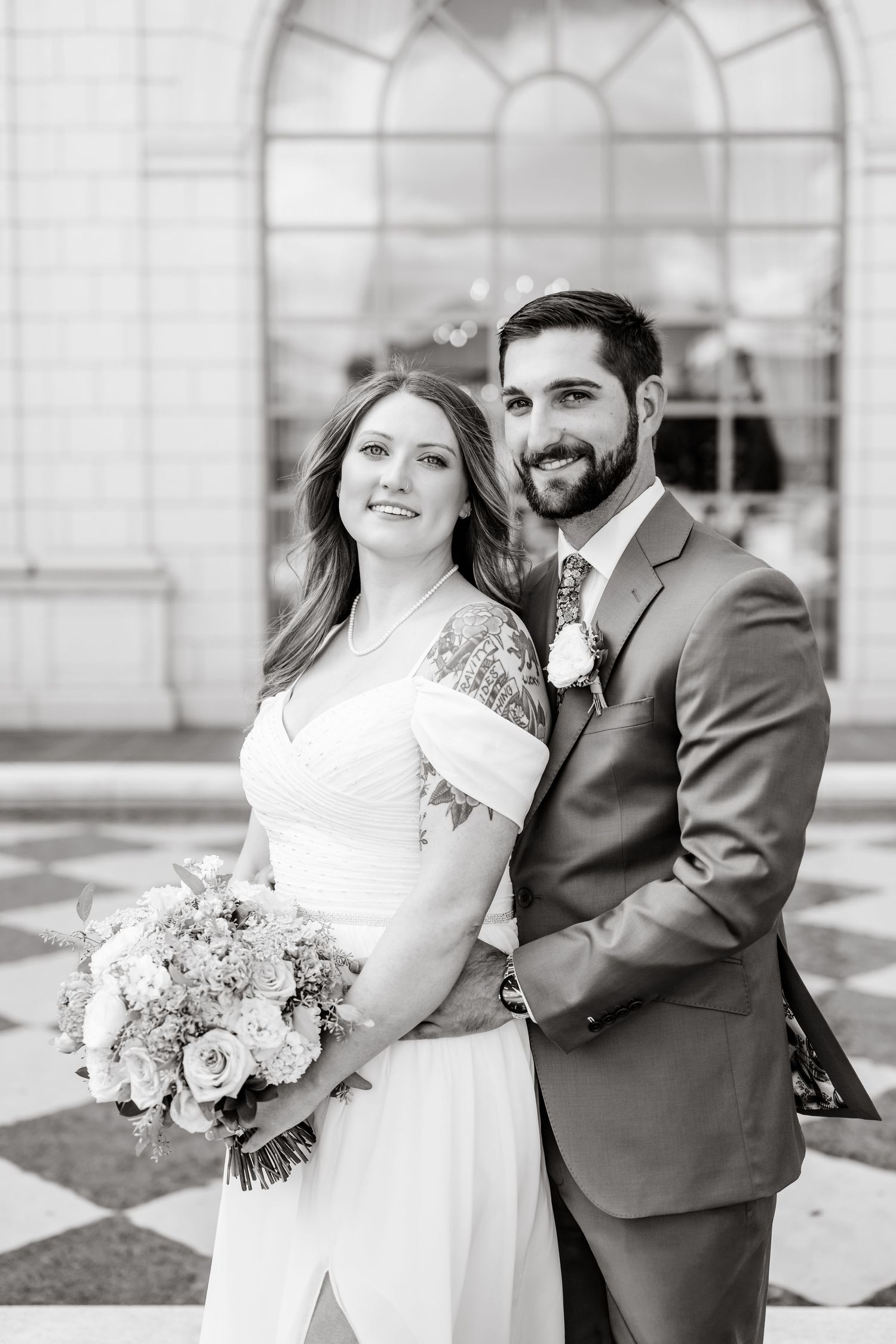 A couple poses together in formal attire with a floral bouquet against an arched window backdrop.