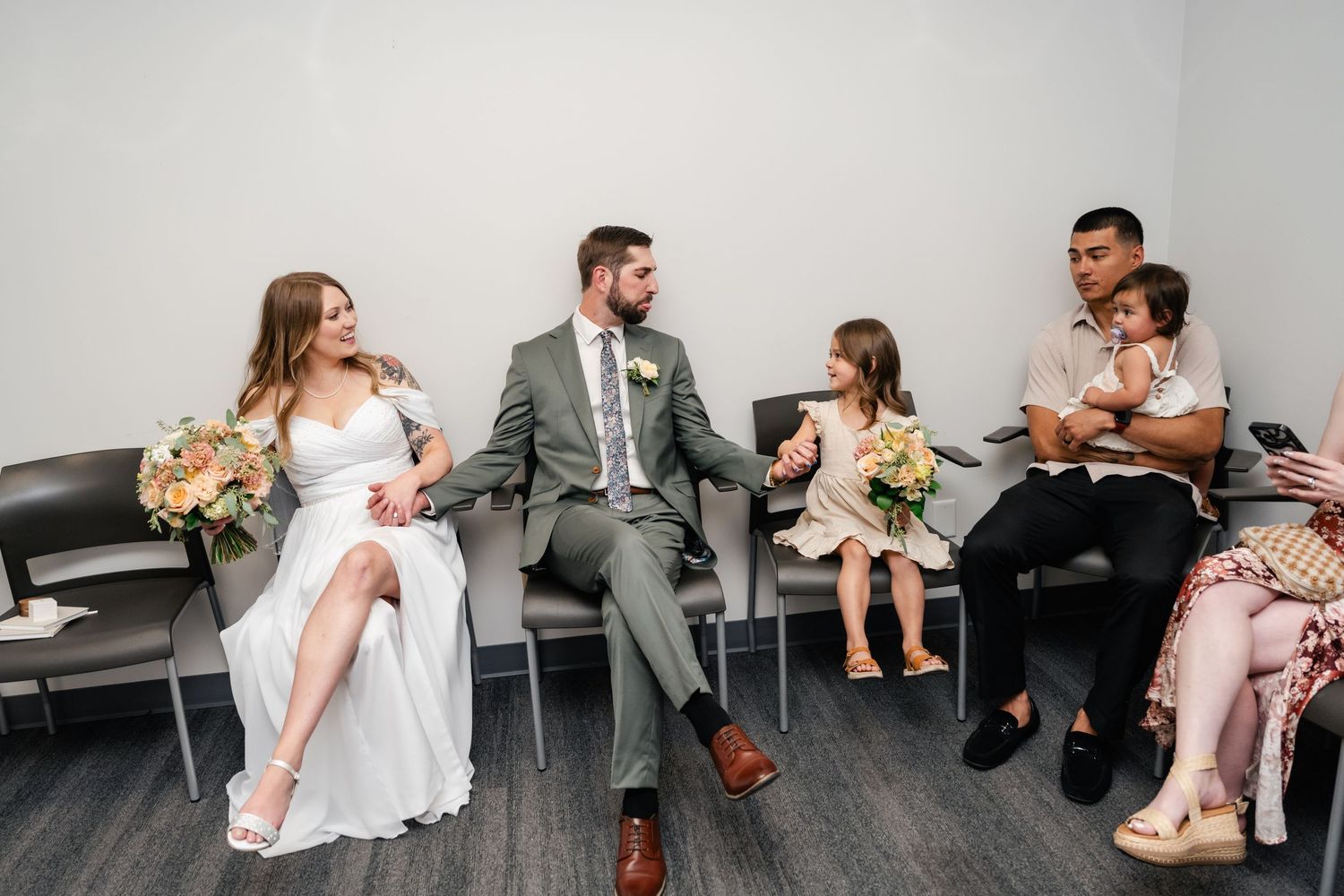 Wedding party relaxes in chairs along a wall during a casual moment at the ceremony.