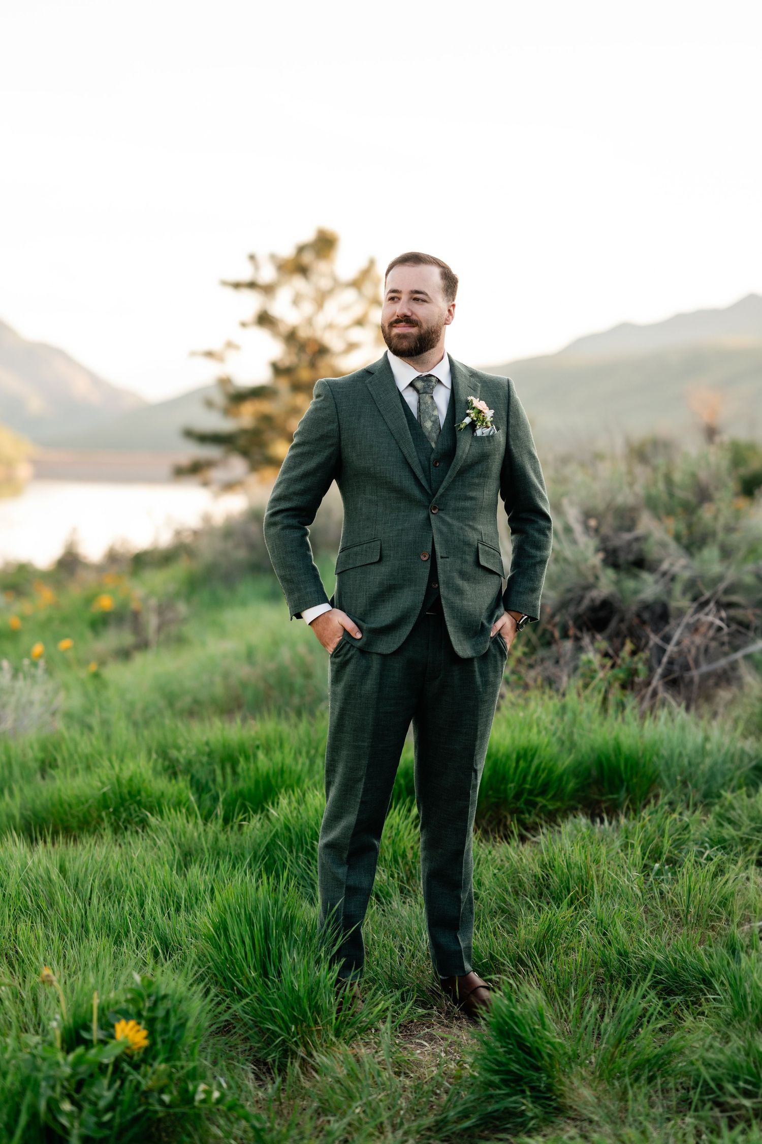 Groom in dark green suit poses in a scenic mountain meadow with wildflowers. Mountain Wedding Photographer.