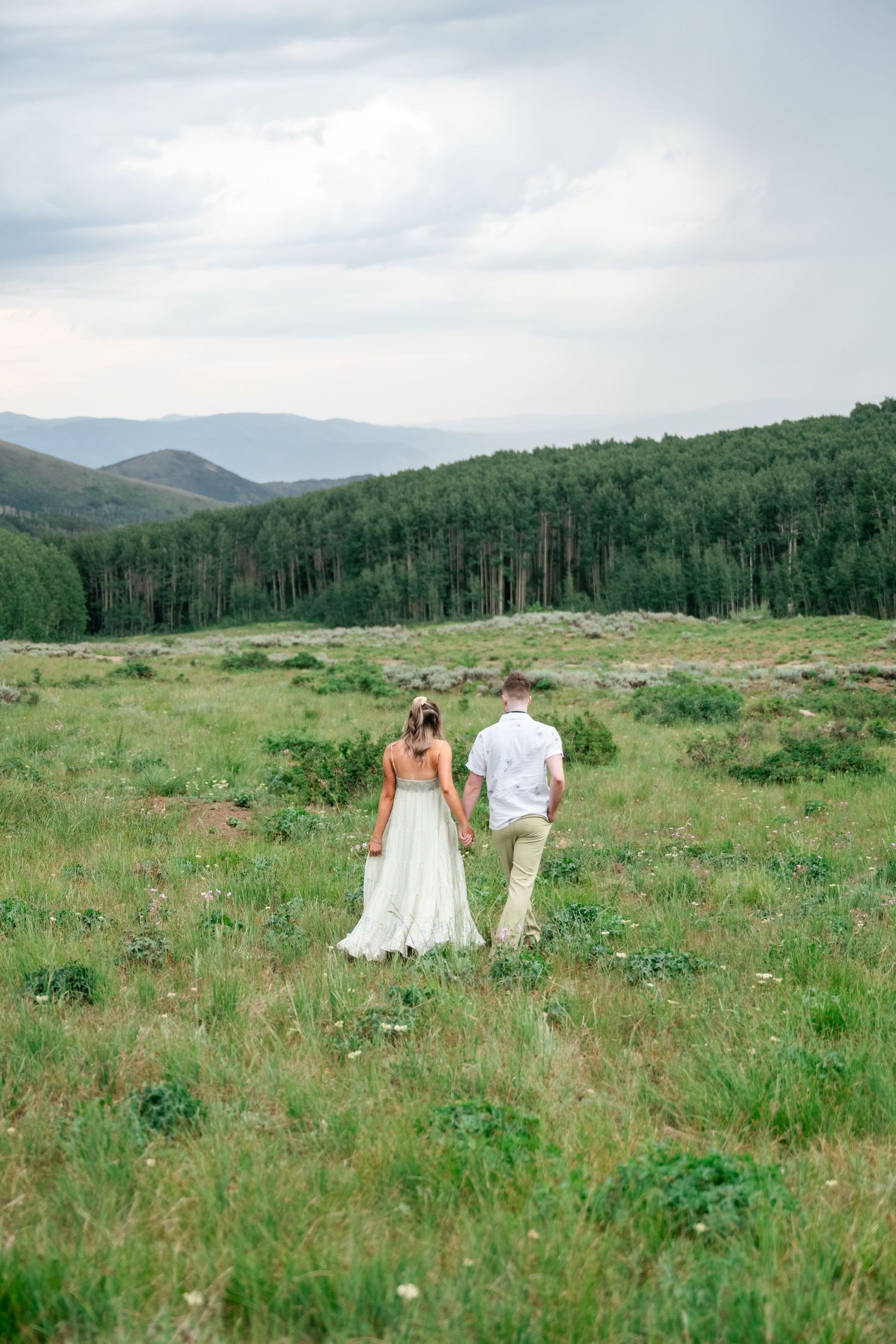 A couple walks hand in hand through a mountain meadow with rolling hills and forest in the distance.