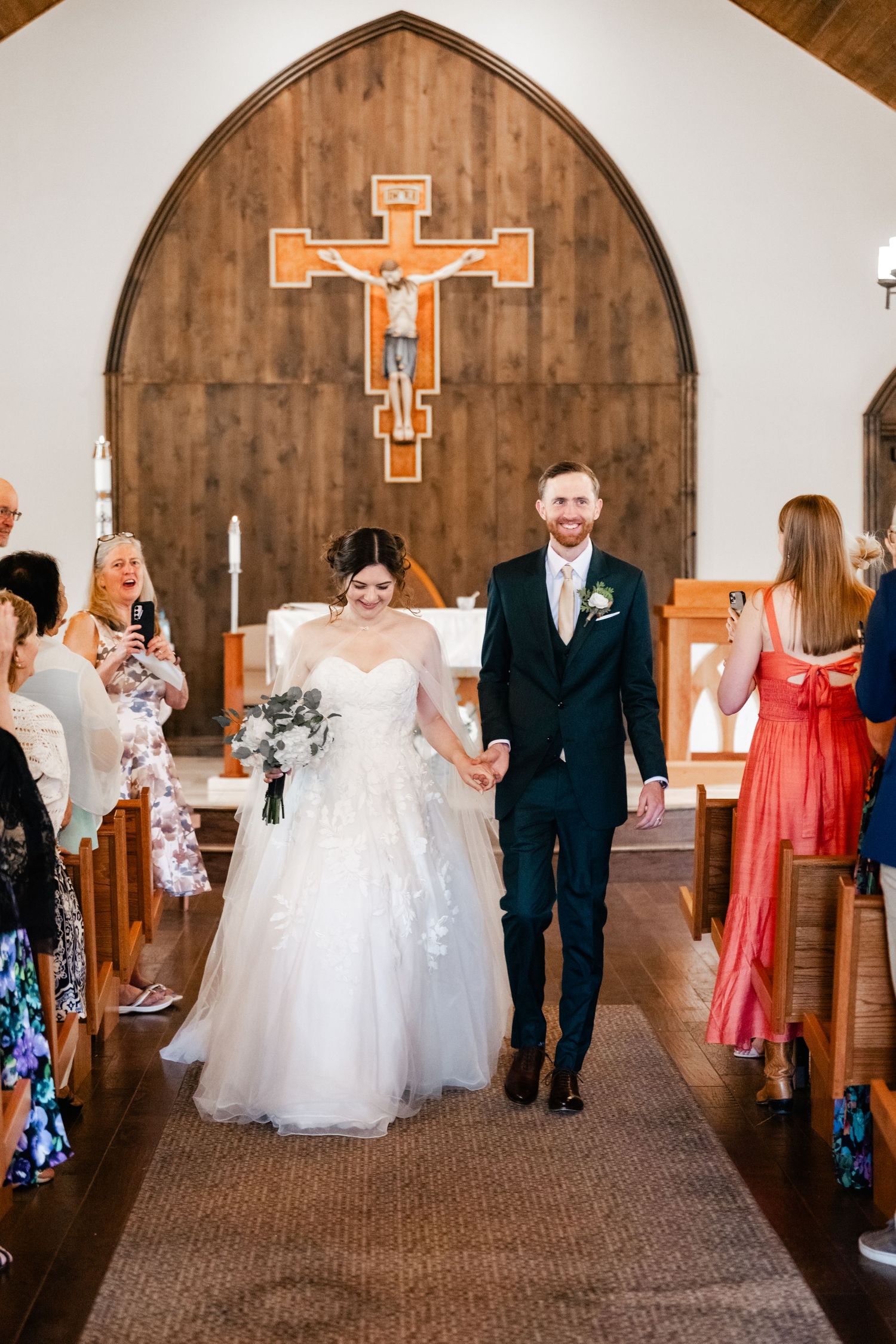 Wedding party processional walking down the aisle in a church with wooden cross and barrel wall decor.