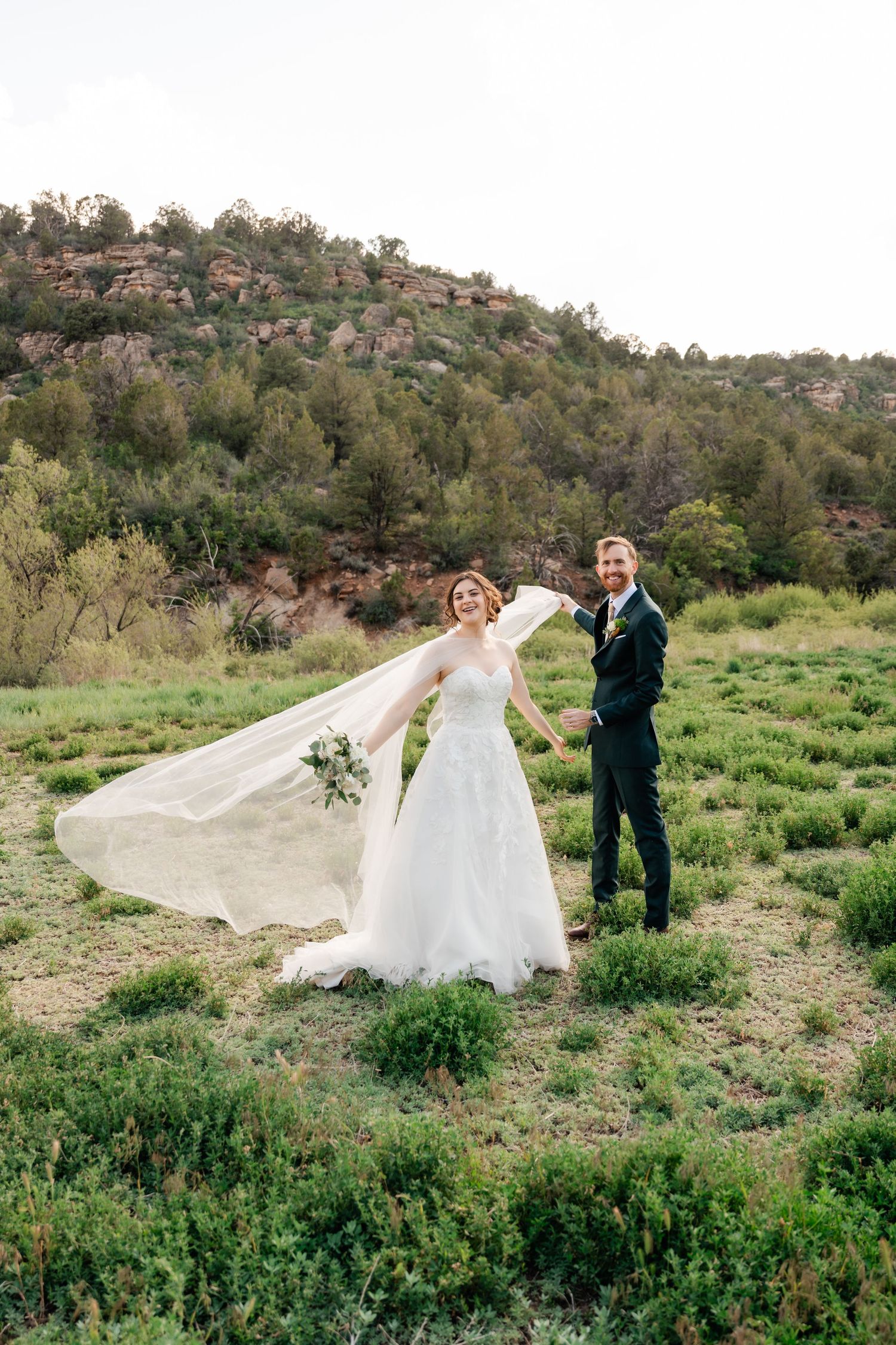A bride's white dress flows in the wind as she walks along a desert path with greenery and rocky hills.