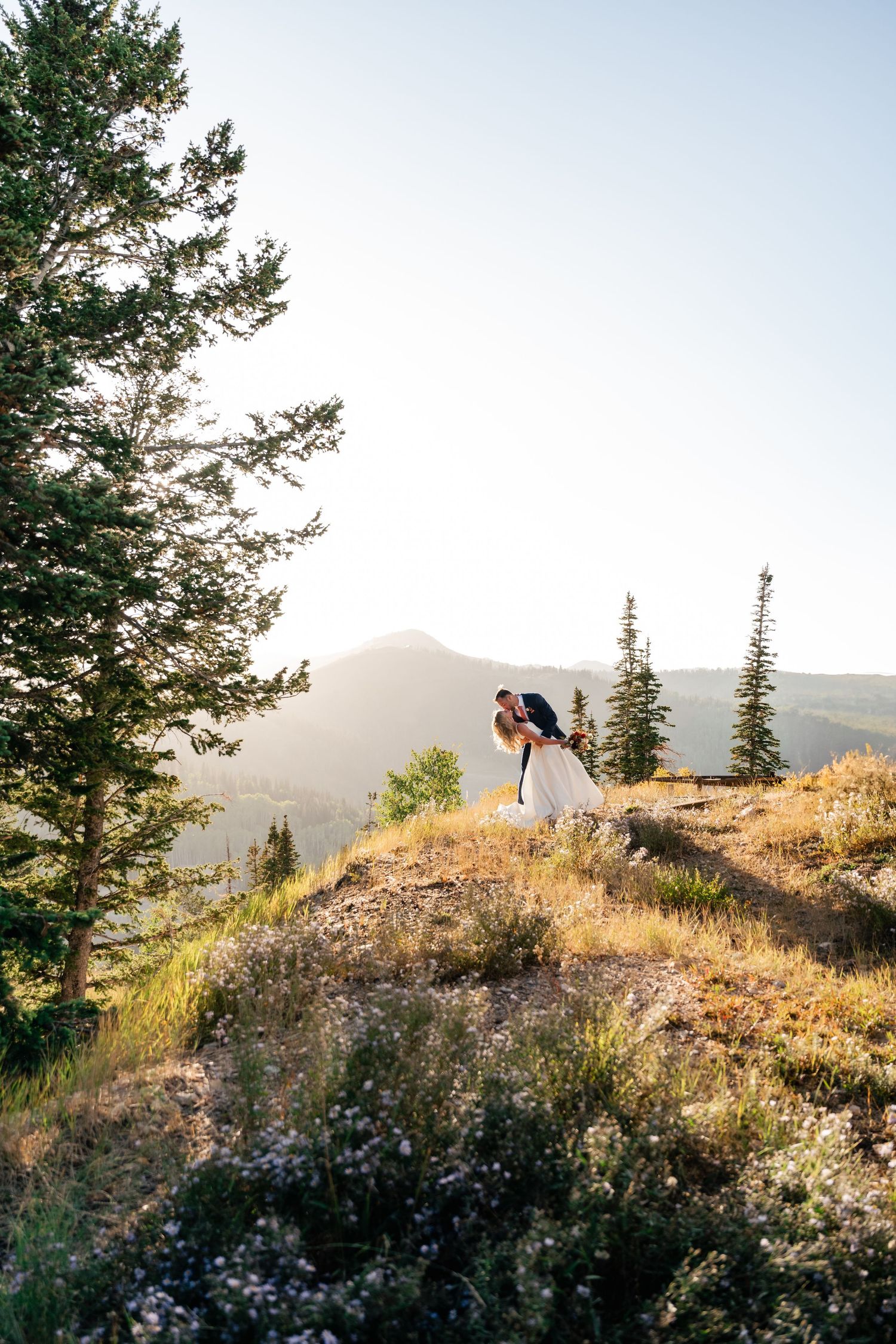 A couple poses on a scenic mountaintop during golden hour with evergreen trees and mountains in the background.