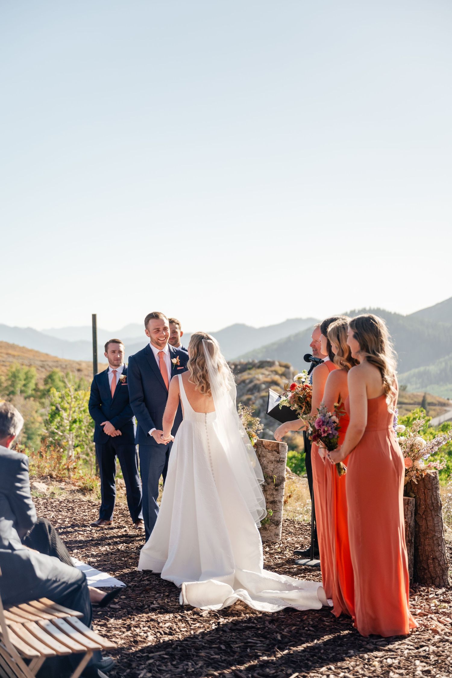 Wedding party in coral dresses and navy suits gather for outdoor ceremony against mountain backdrop.