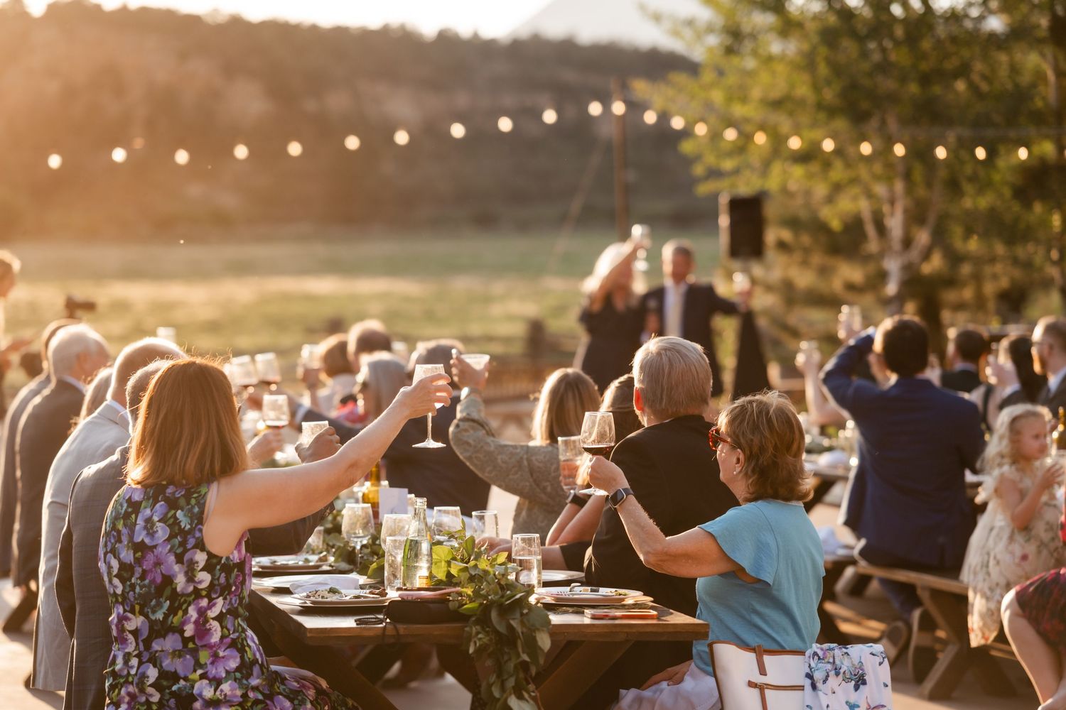 Wedding guests toast during golden hour under string lights at an outdoor reception.