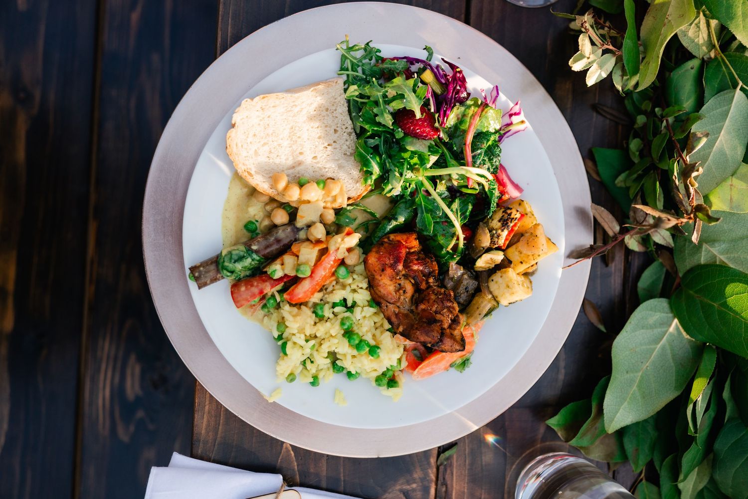 Elegant plated dishes of colorful salads and entrees arranged in a row on a dark wooden surface with green foliage.