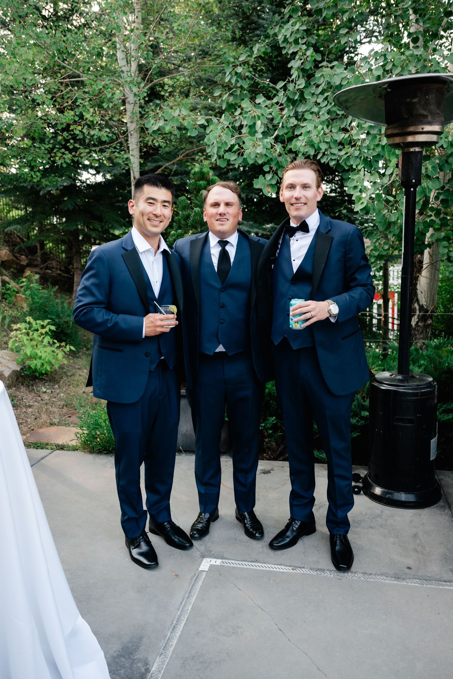 Groomsmen in navy suits gather outdoors for drinks during wedding celebration.