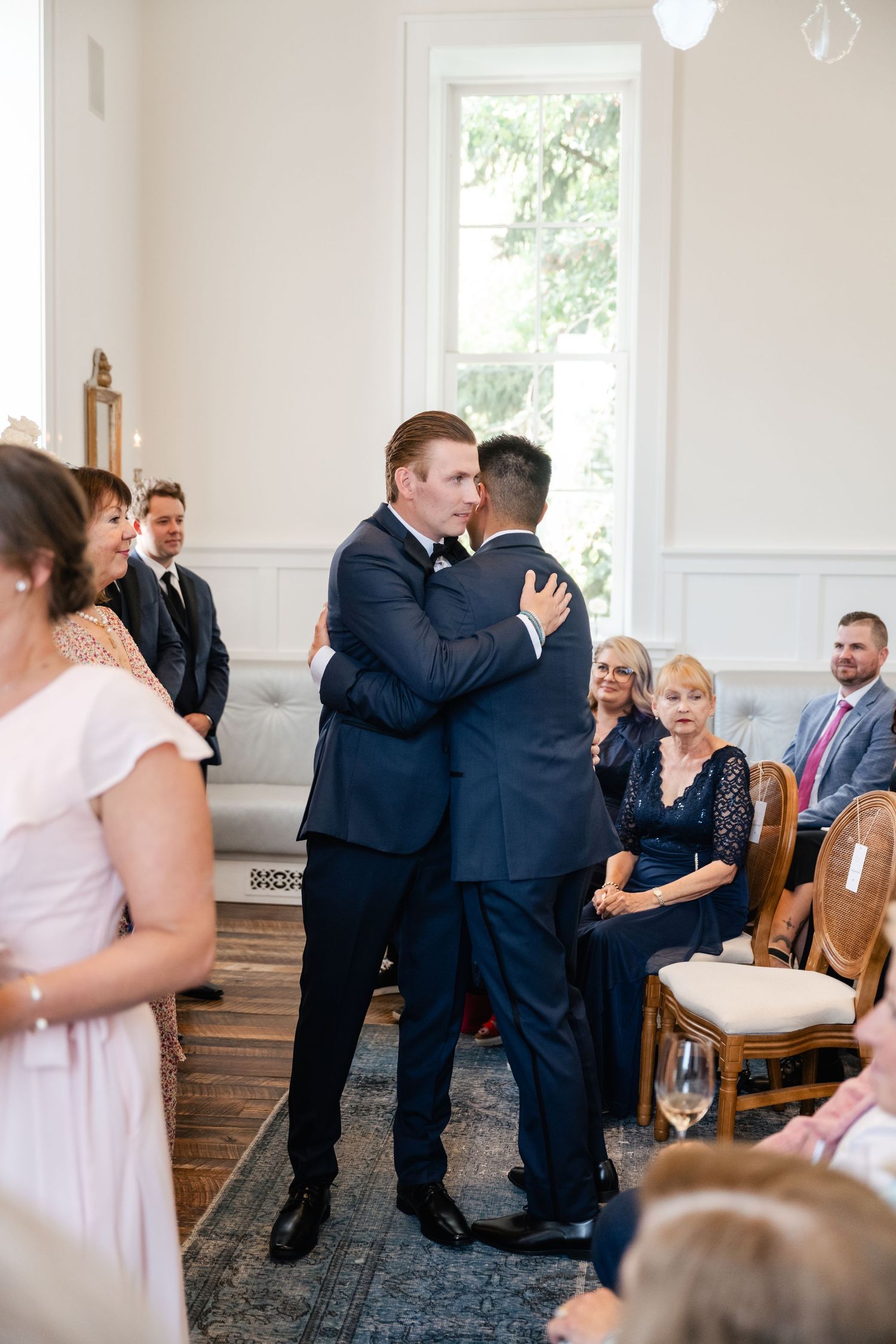 Wedding ceremony guests mingle and embrace in a bright indoor venue with wooden chairs.
