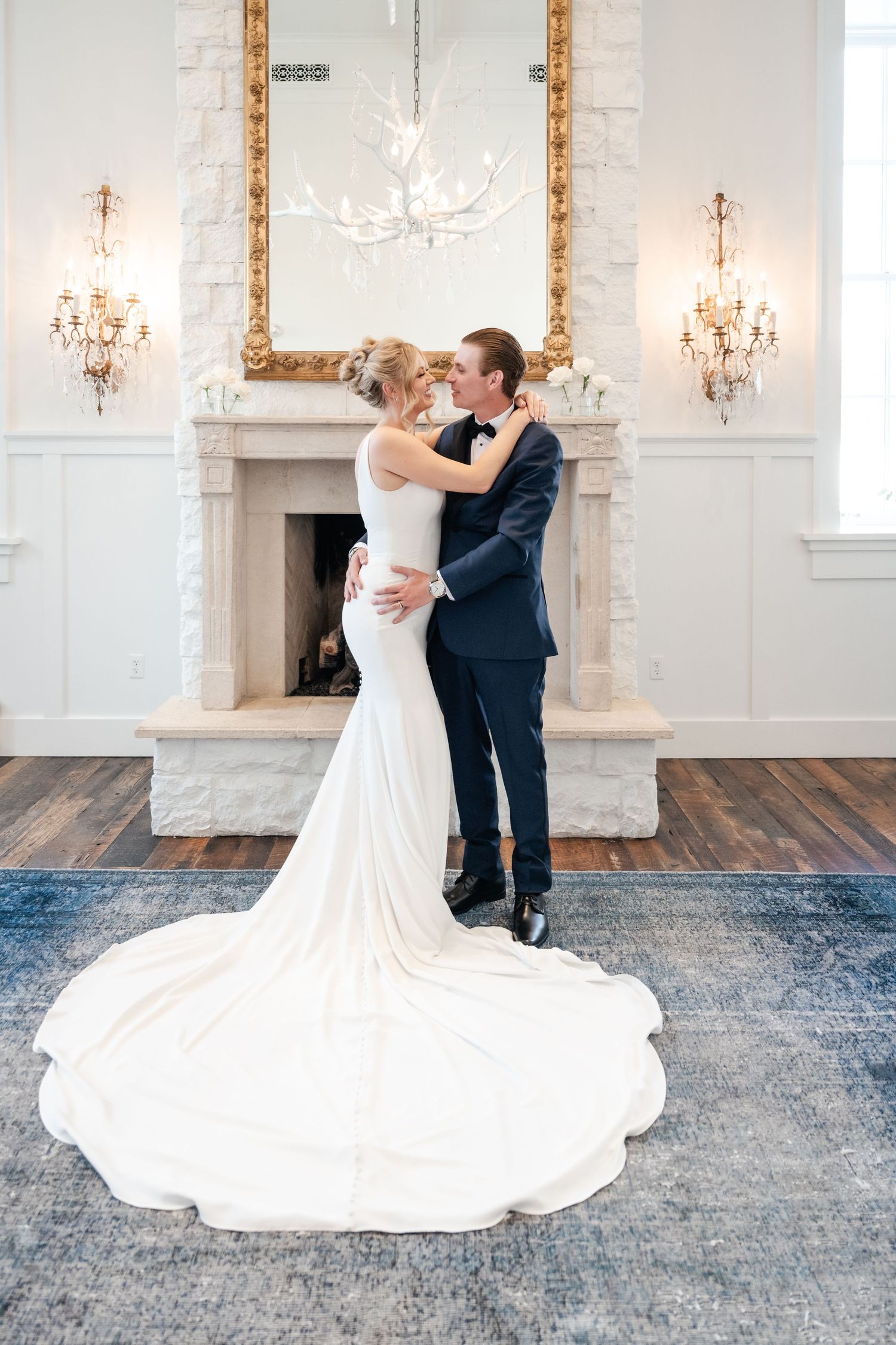Elegant wedding couple embraces in front of ornate fireplace with crystal chandeliers and gold mirror.