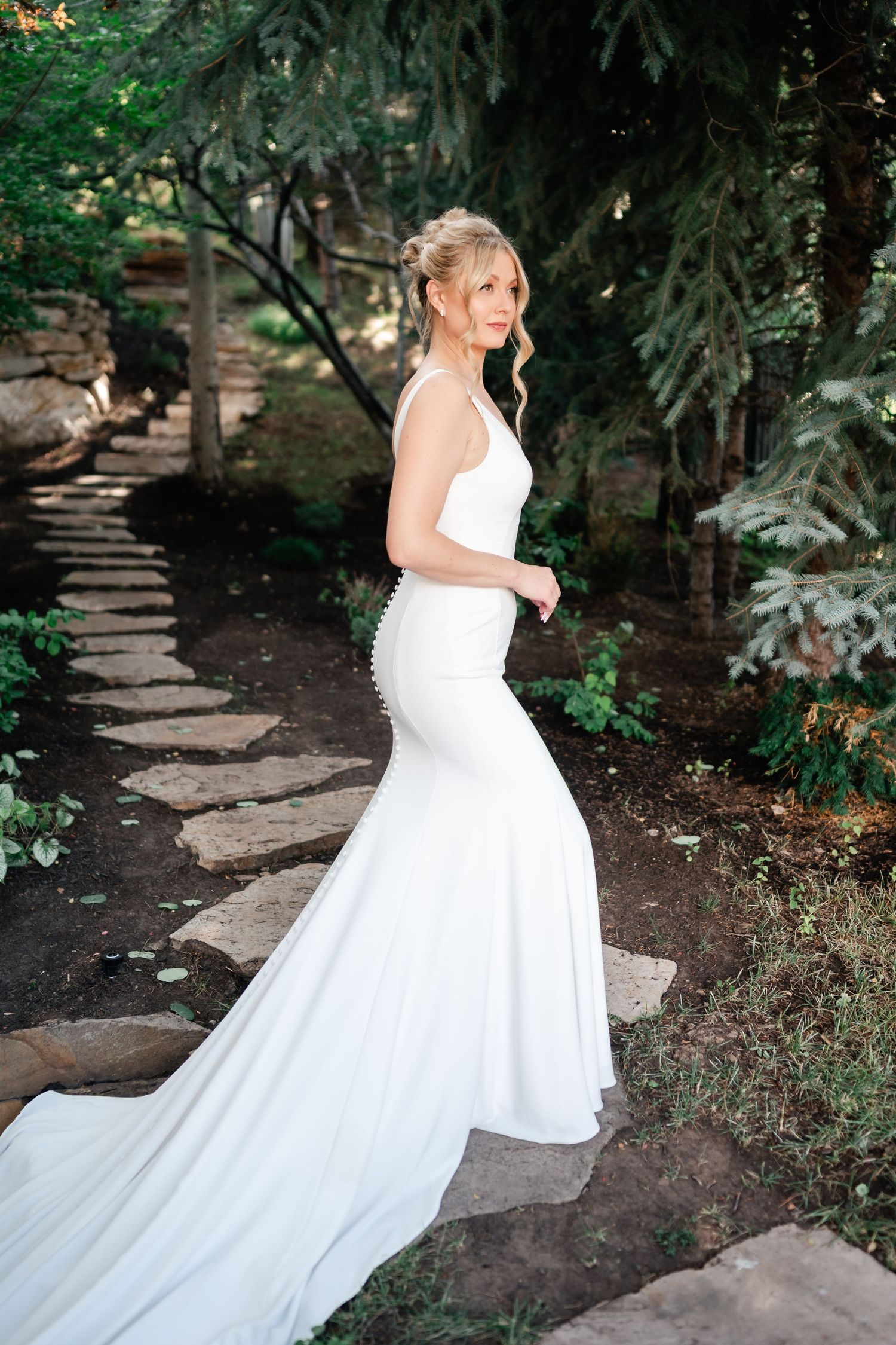 Bride in flowing white gown walks along stone garden path surrounded by evergreens.