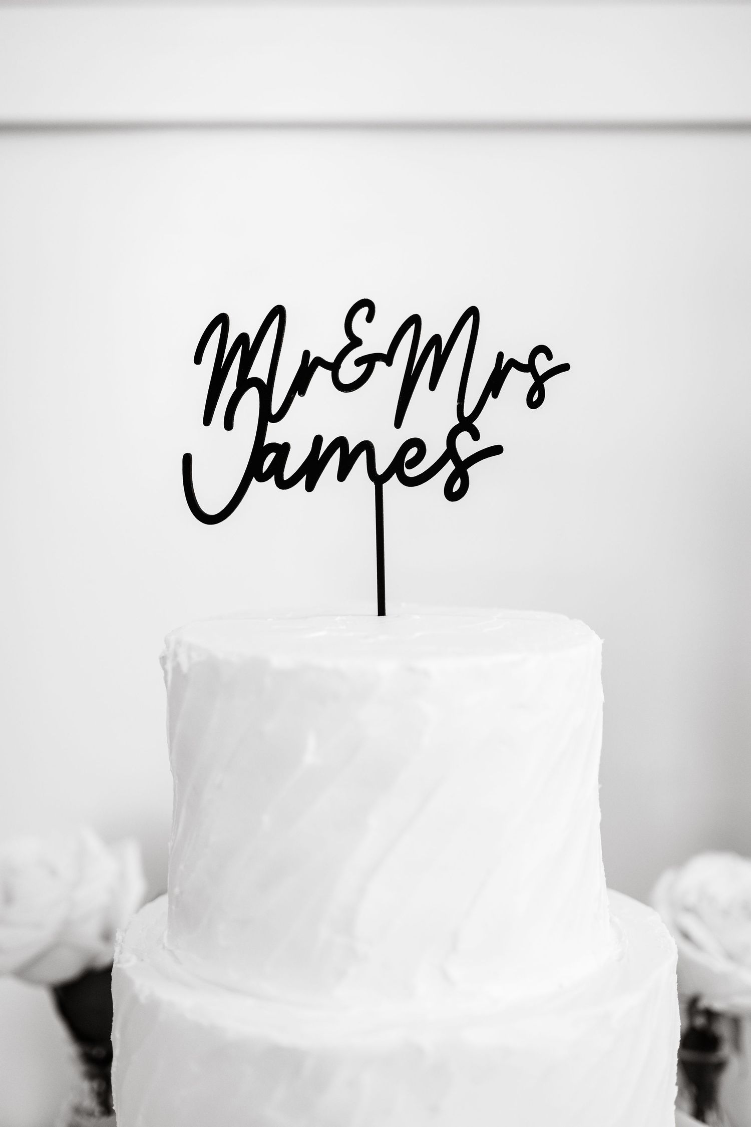Black Mr & Mrs James cake topper on a white two-tier wedding cake with white roses in background.