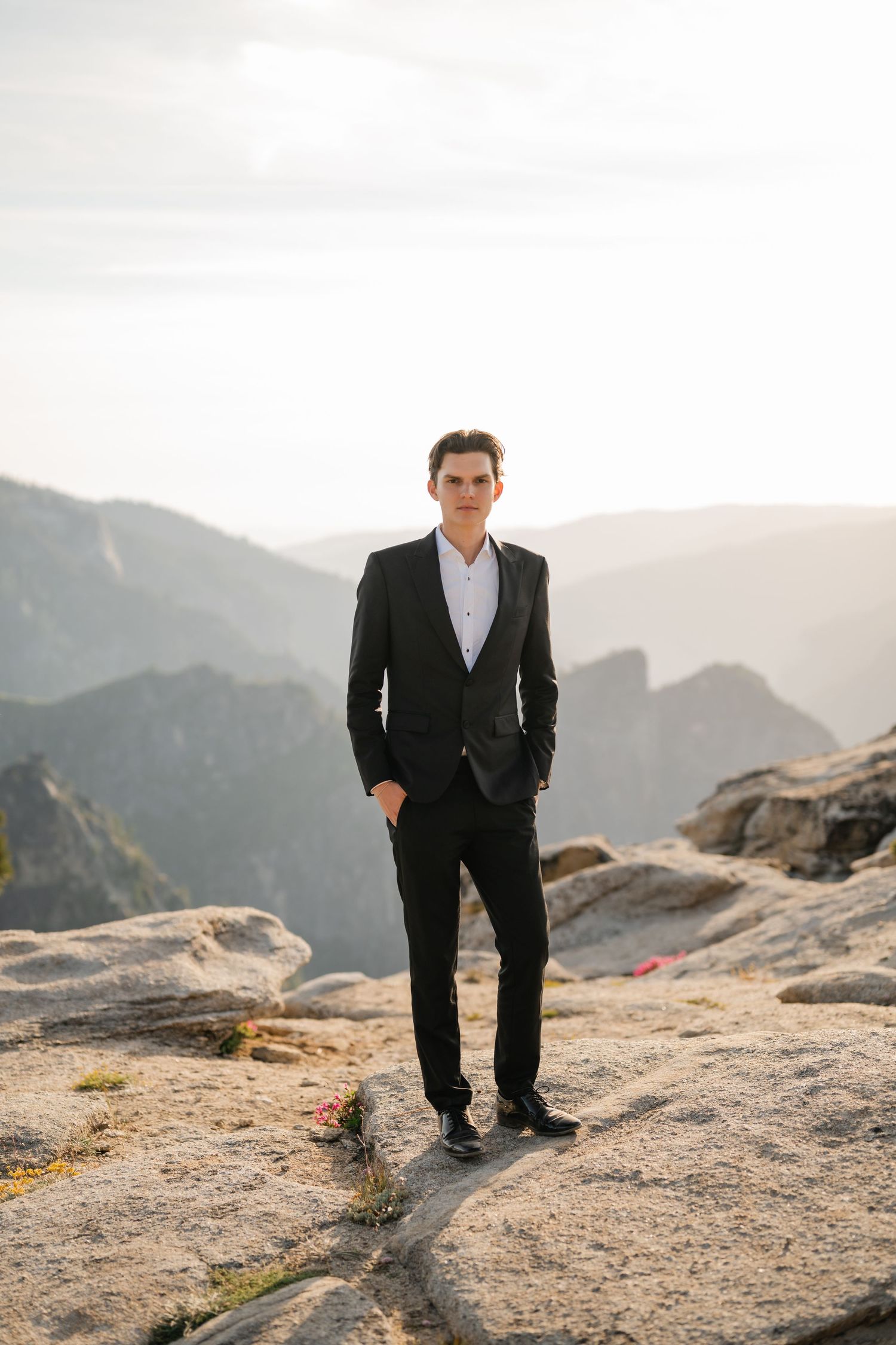 Groom in black suit stands confidently on rocky mountain terrain with misty valley backdrop.