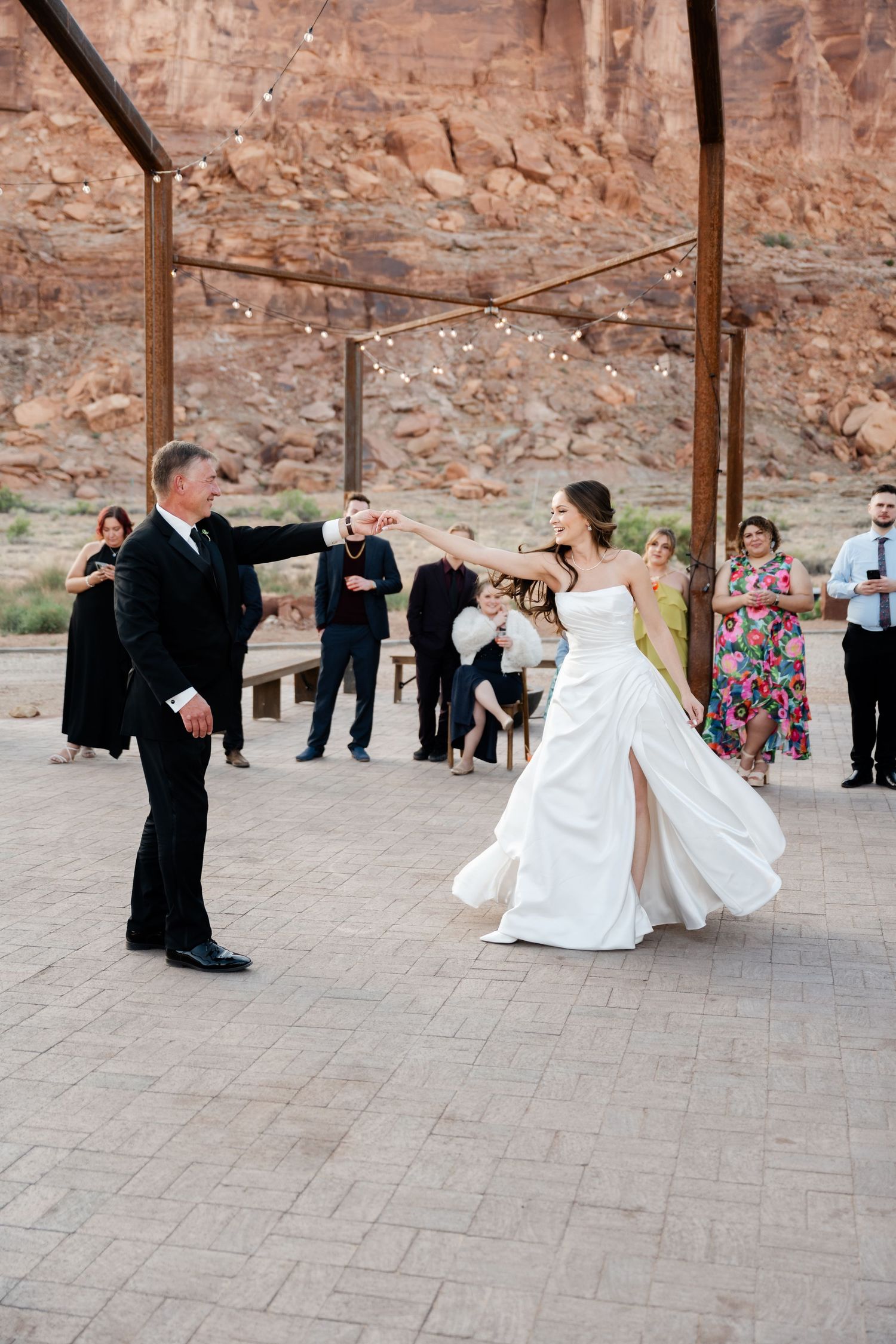 Bride and groom share first dance outdoors at desert wedding venue with red rock canyon backdrop and string lights.