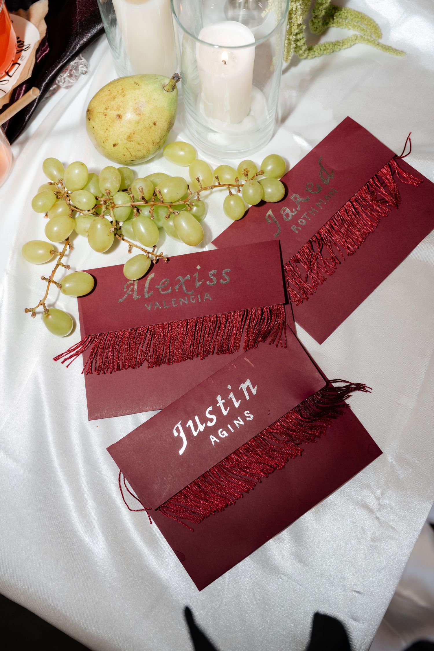 Elegant burgundy place cards with fringe detail for Alexiss Valencia and Justin Agins, displayed with grapes and a pear on white linen.