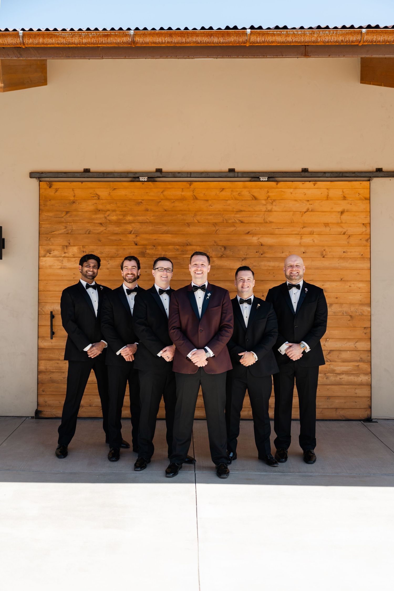 Groom in burgundy tuxedo stands centered with five groomsmen in black tuxedos before rustic wooden barn doors.