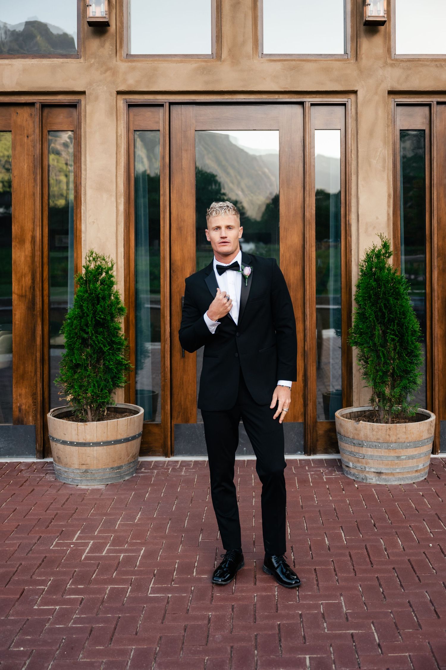 Classic formal attire against wooden doorway with potted cypress plants and mountain backdrop.
