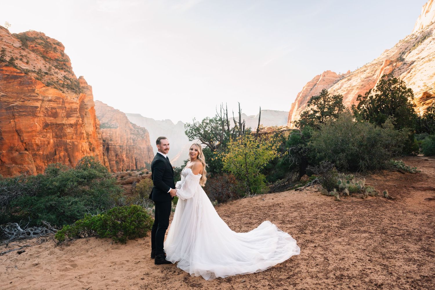 Wedding couple poses on desert trail with dramatic orange canyon walls in background.