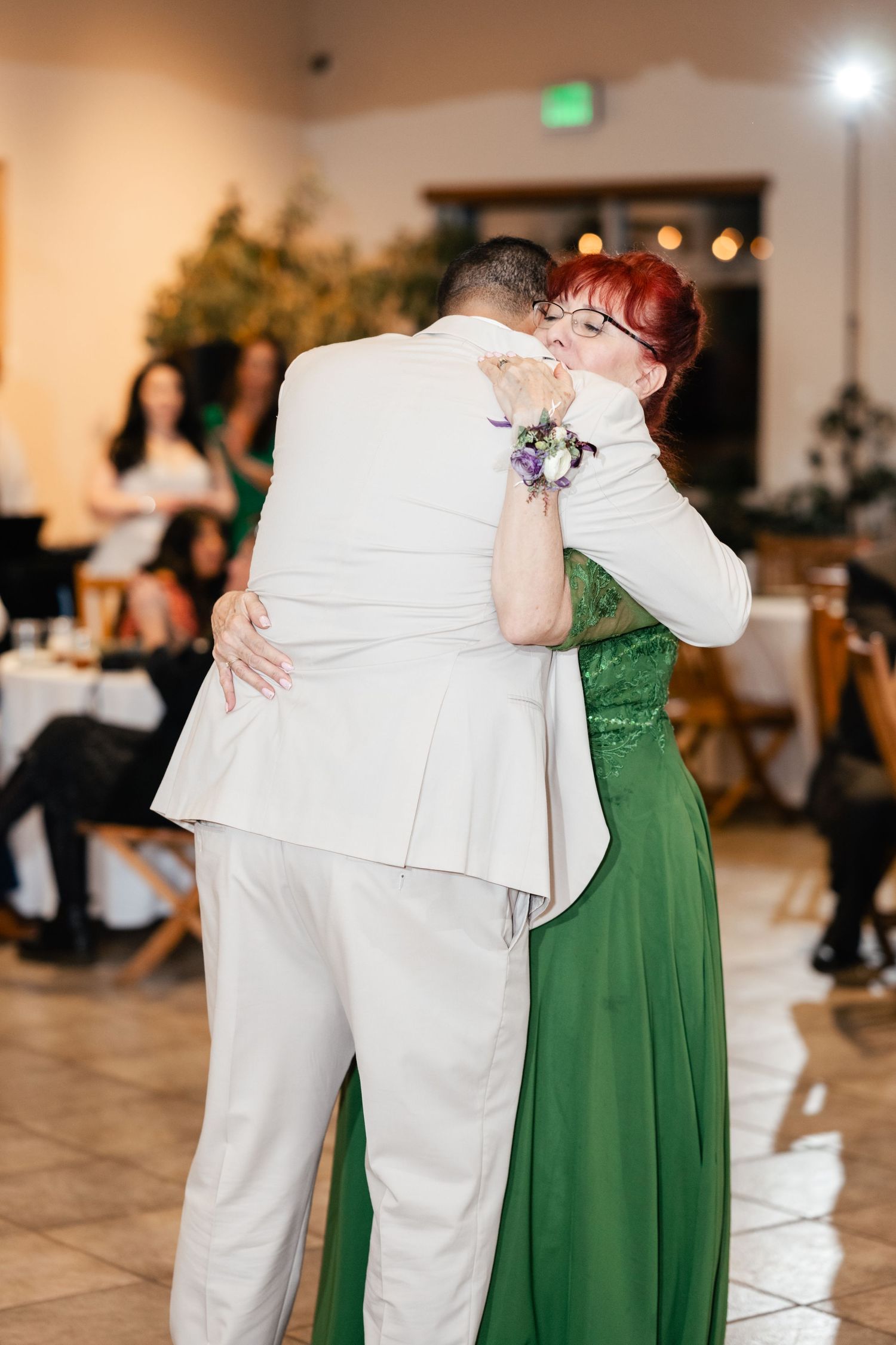 Two people embrace on the dance floor, one wearing a white suit and the other in a flowing green evening dress.